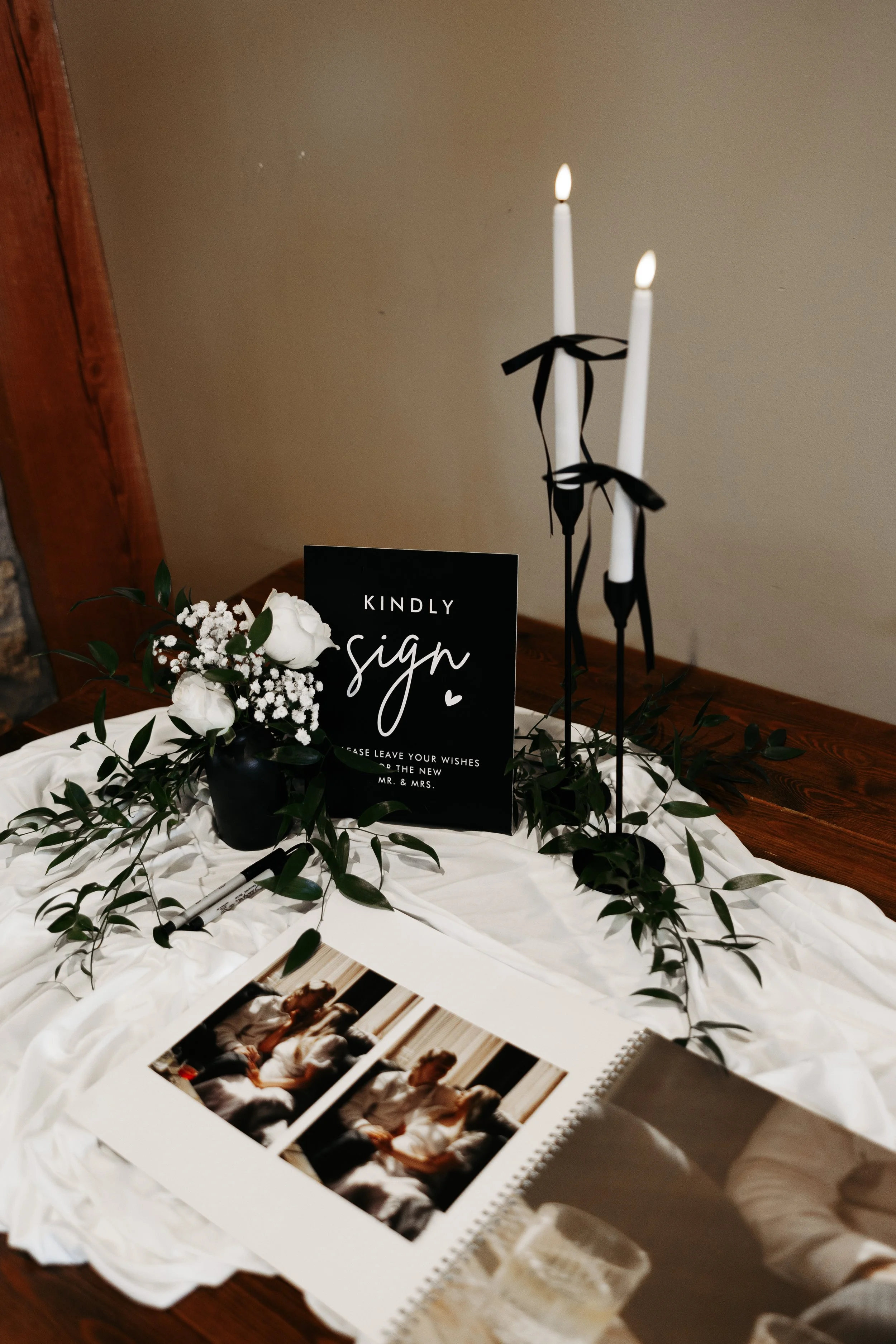Wedding or event table setting with white candles, a black sign with white lettering that reads 'Kindly sign', a floral arrangement of white roses, greenery, and baby's breath, a pen, photos of a couple, and a glass of champagne on a white cloth on a