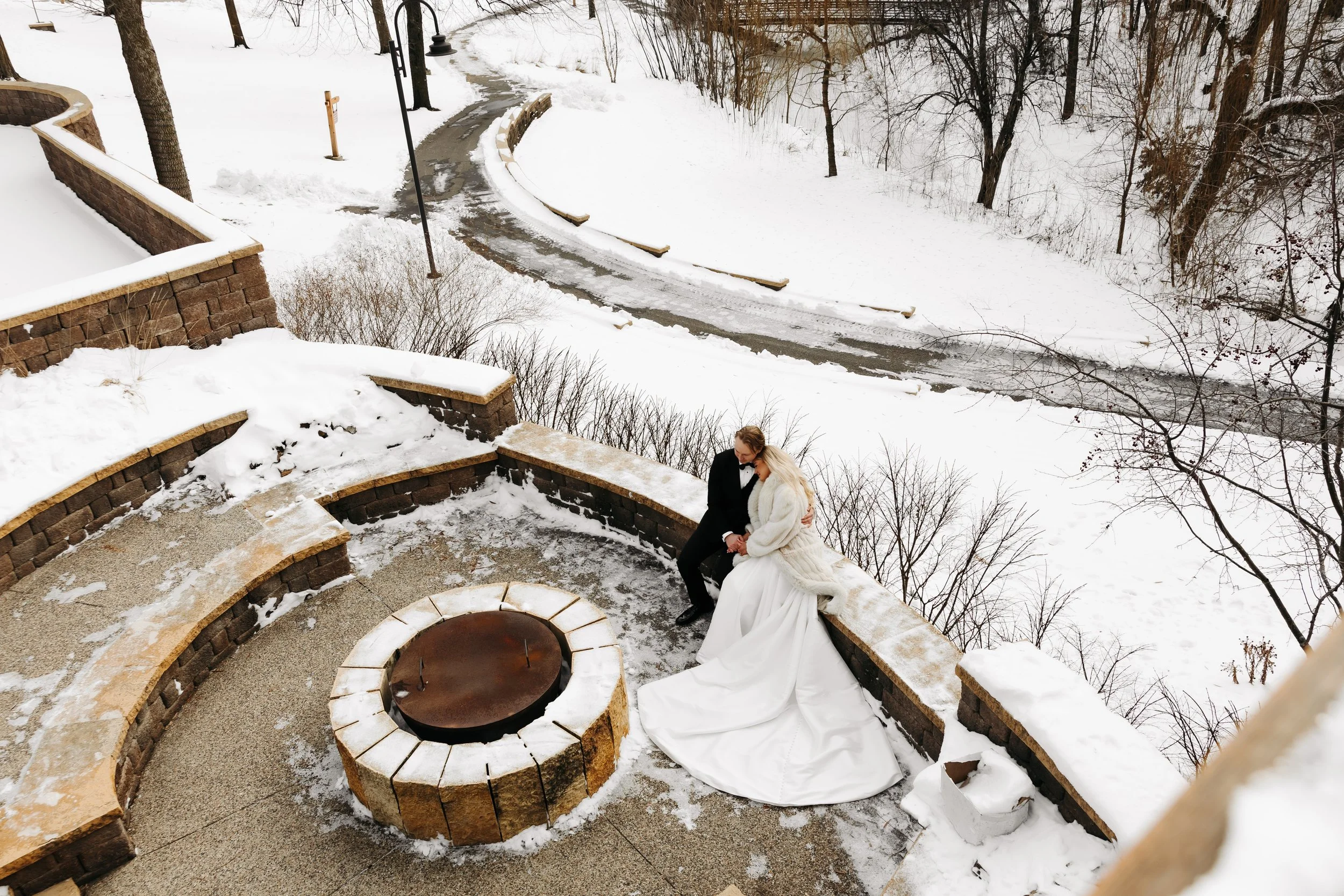 A bride and groom sitting close together on a snow-covered outdoor stone bench near a fire pit, with snow and trees in the background.
