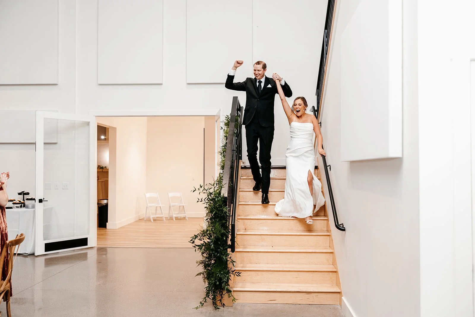 A newlywed couple celebrating as they walk down a staircase after their wedding, with the groom in a dark suit lifting the bride's hand in victory.