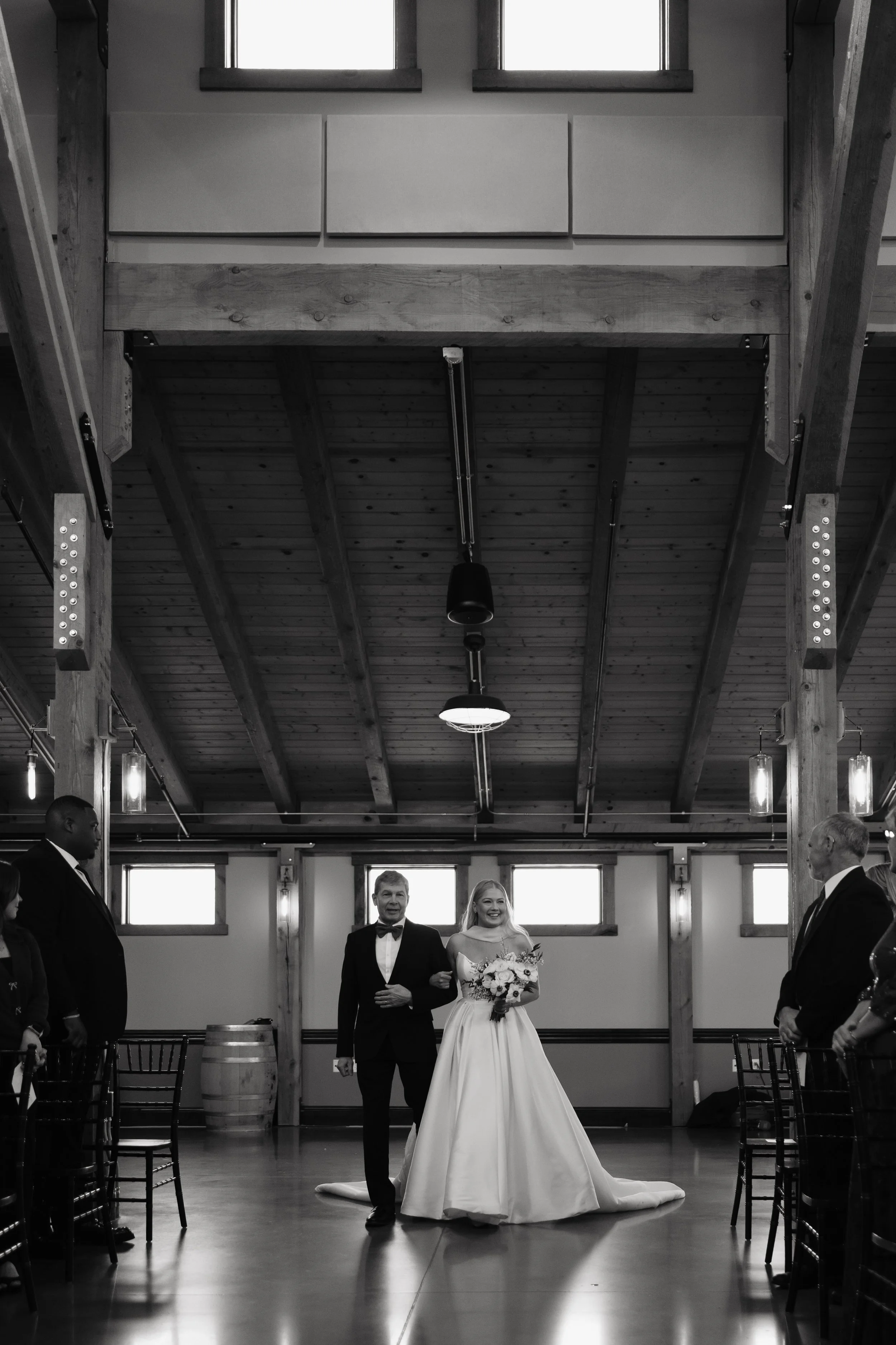 Bride walking down the aisle with her father at her wedding venue with guests on either side, in a rustic wooden hall.
