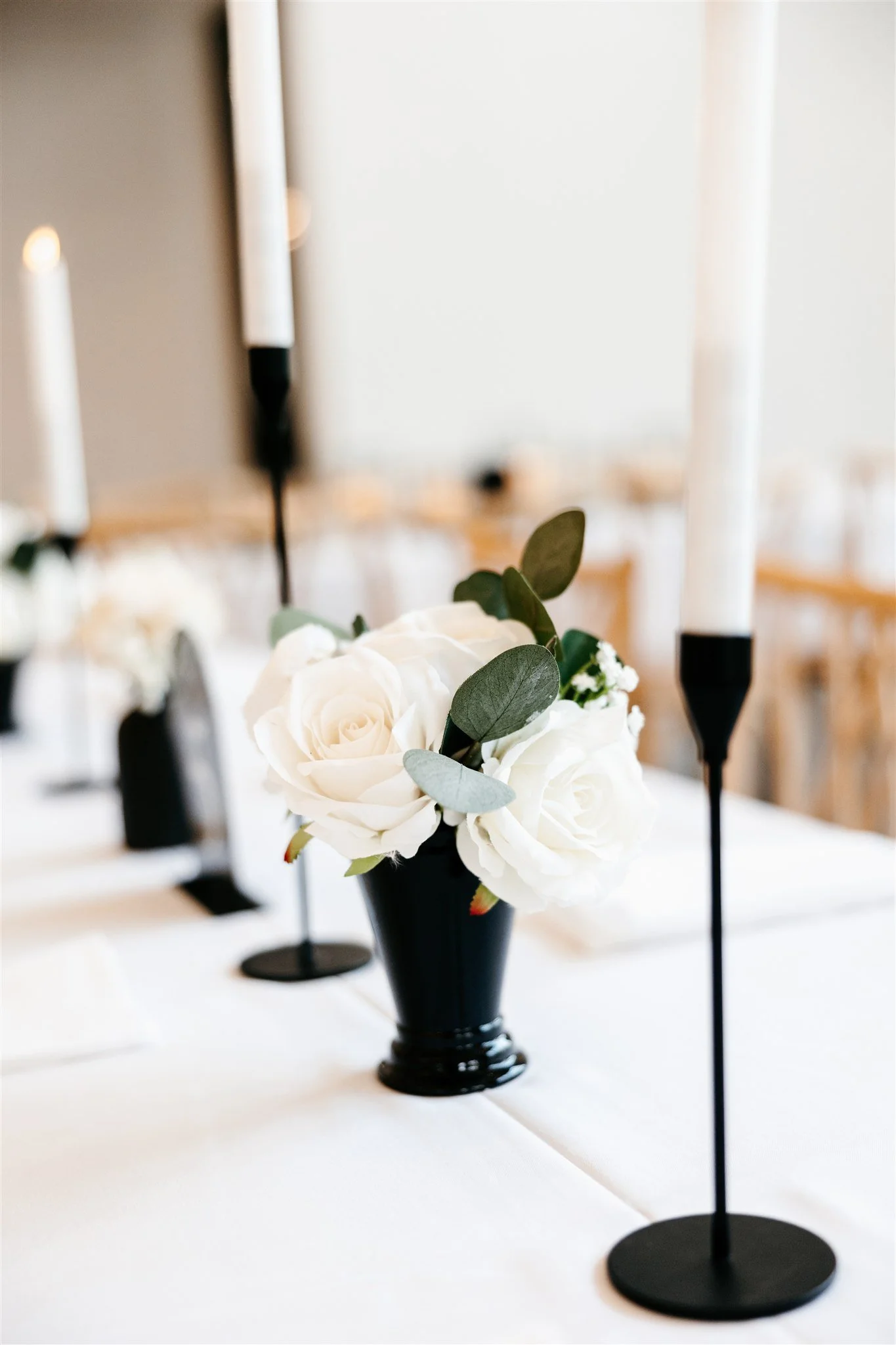 Close-up of a floral centerpiece with white roses and green leaves in a black vase on a white table, flanked by tall white candles in black holders.