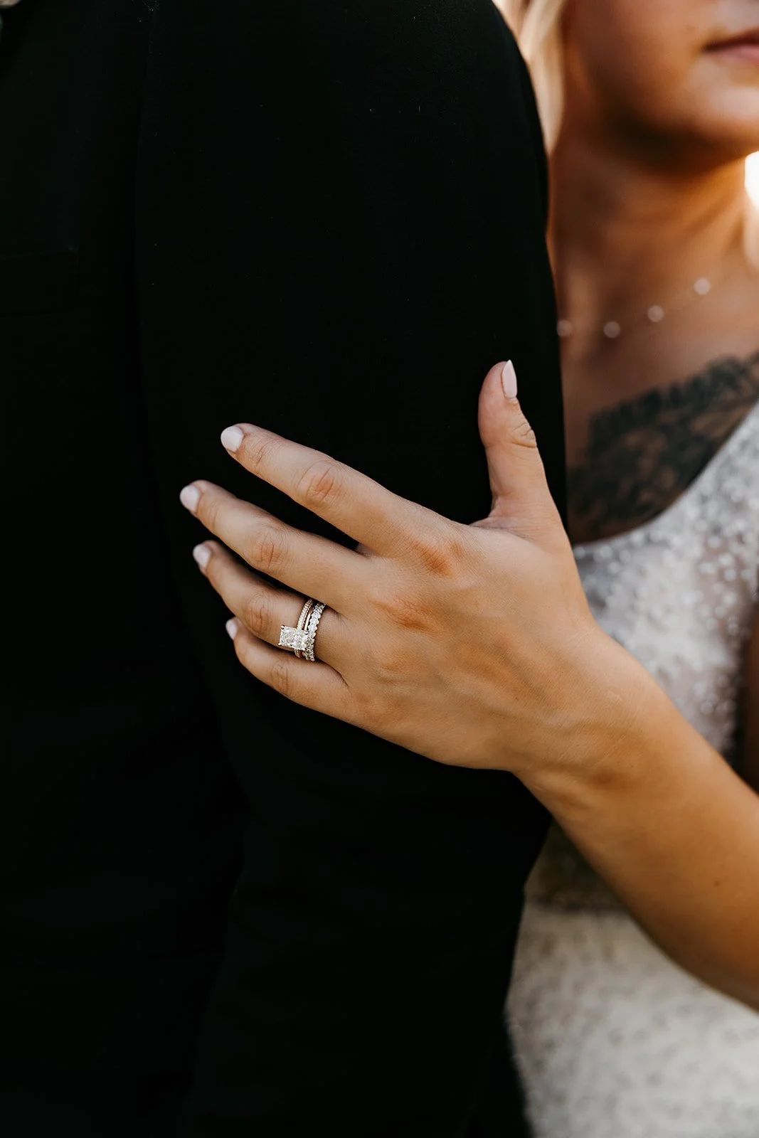 A close-up of a woman's hand wearing a wedding ring on her ring finger, resting on a man's arm dressed in black, with a woman in a white dress visible in the background.