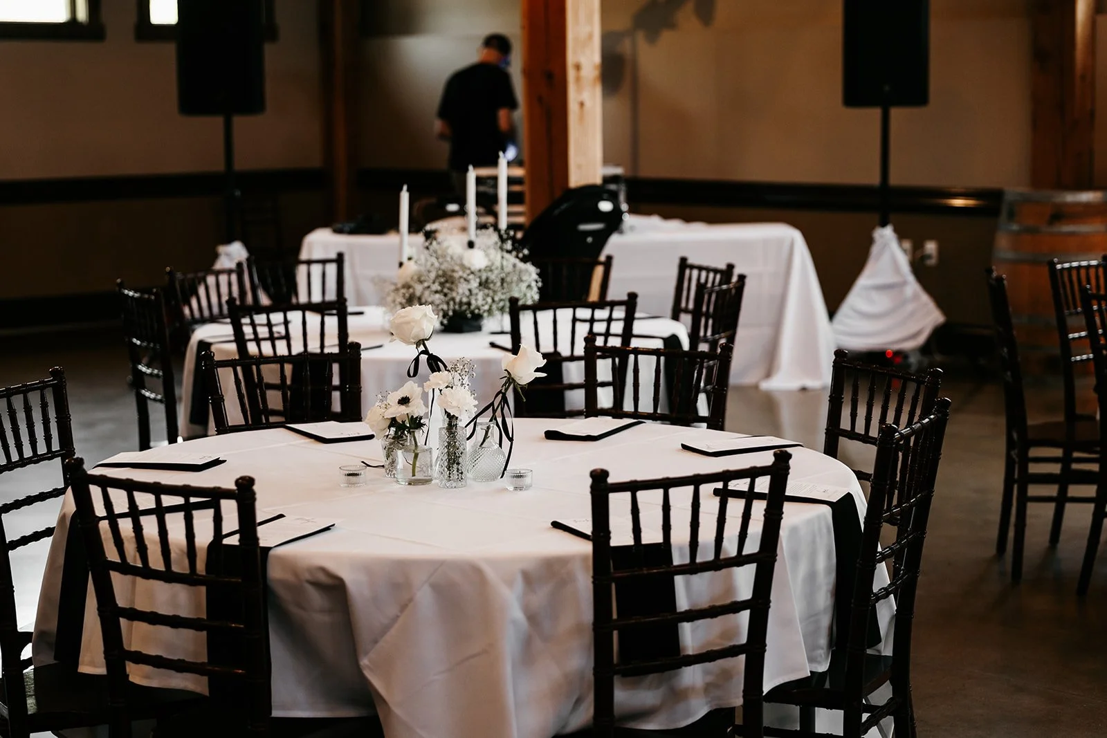 Wedding reception setup with round tables covered in white tablecloths, decorated with white floral centerpieces and black chairs, in a rustic venue with wooden beams.