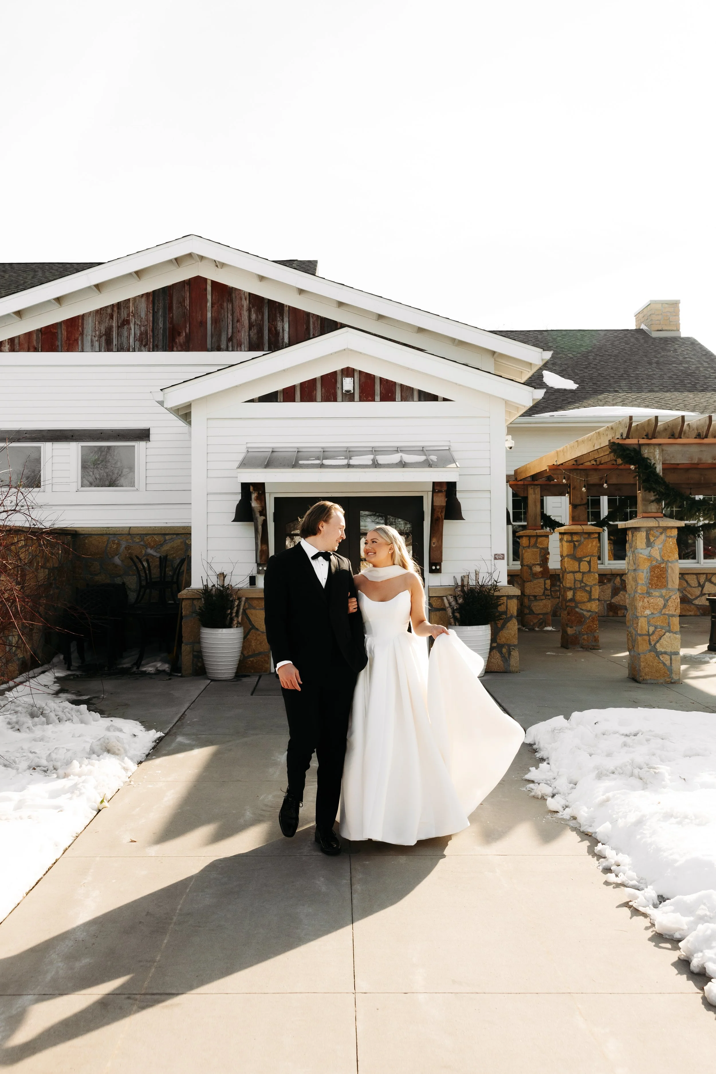 A bride and groom walking outdoors on a snowy day, holding hands and smiling at each other, in front of a rustic white house with a stone foundation.