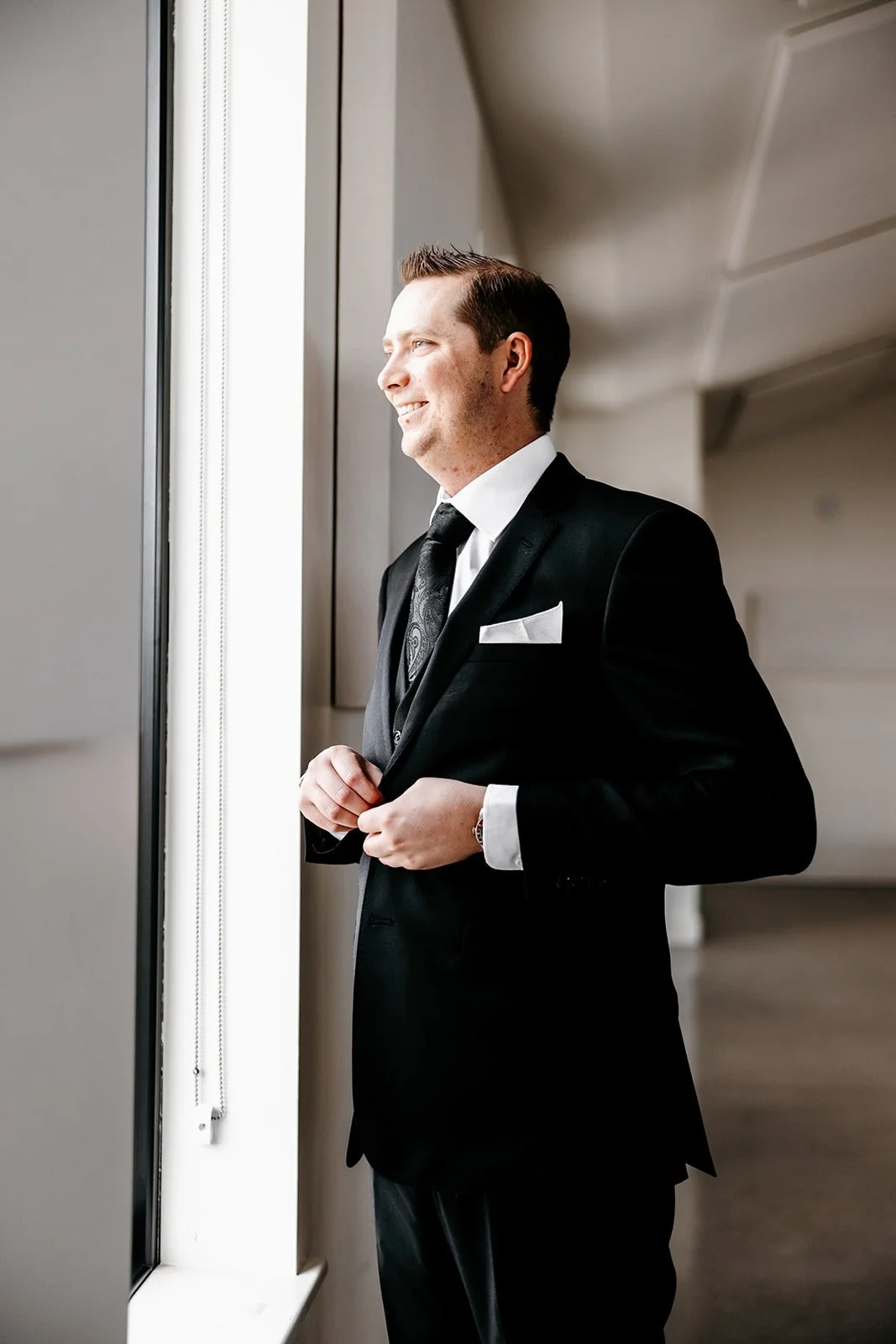 A young man in a black tuxedo, white shirt, and black tie looks out a window, smiling and enjoying the moment.