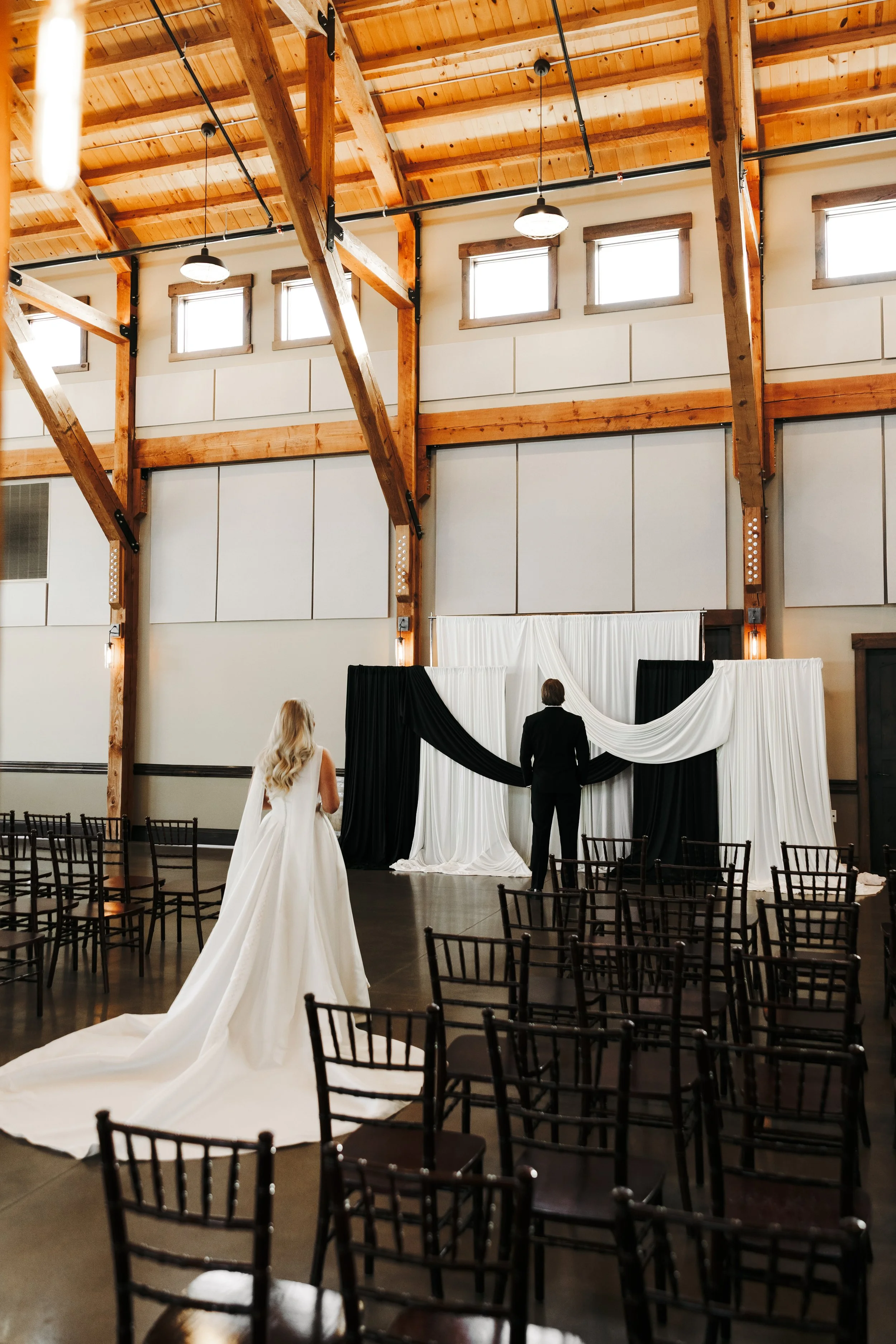 A bride in a white wedding gown and a groom in a black suit standing at an altar with black and white drapes in a rustic indoor wedding venue with wooden beams and chairs arranged for guests.