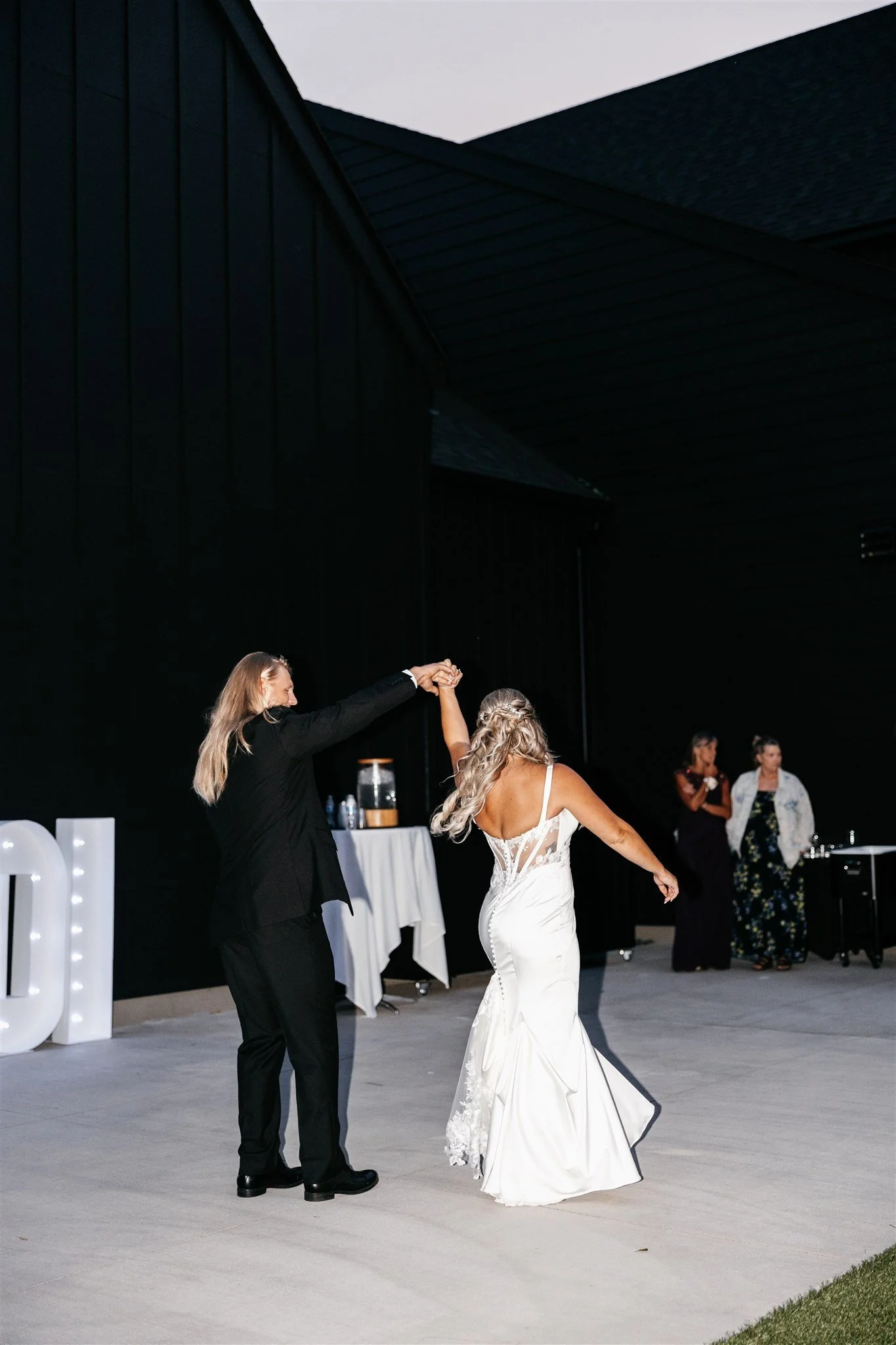 A bride and groom dancing at their wedding reception, with the groom twirling the bride on a dance floor outside a dark building.