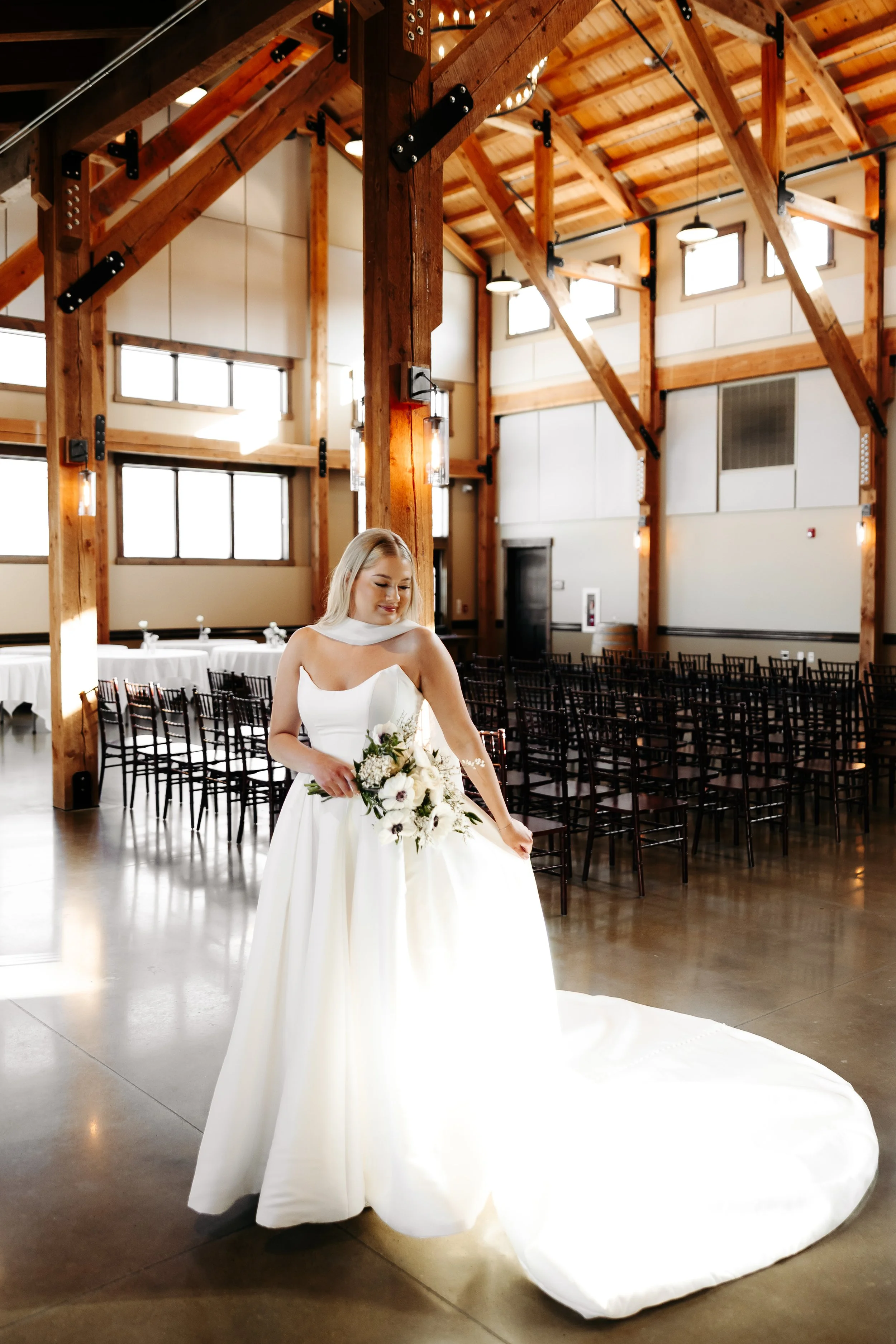 A bride standing with a bouquet in a wooden wedding venue with high ceilings and large windows.