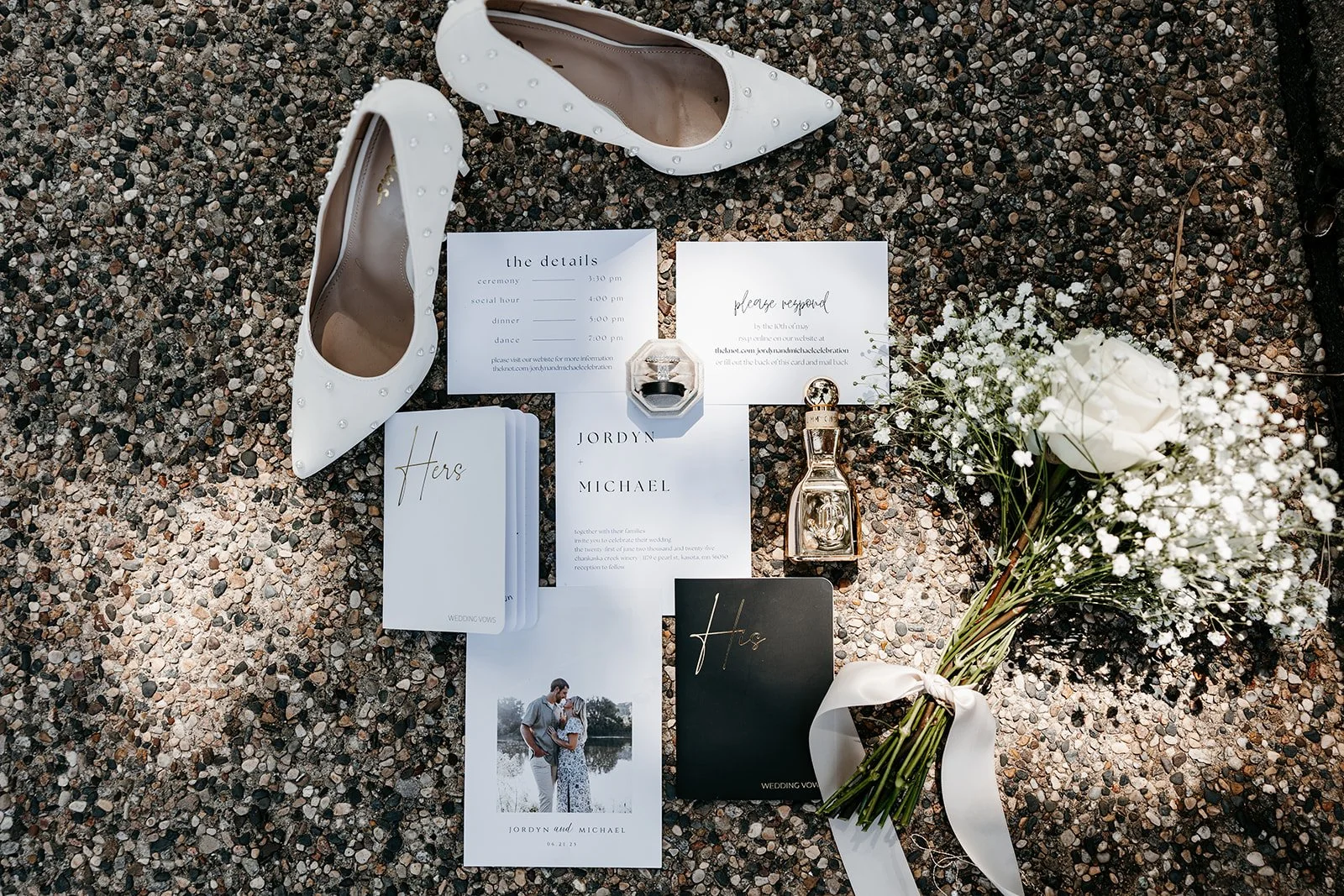 A flat lay of wedding items on gravel ground, including white high-heeled shoes with pearl accents, wedding invitations, a small bouquet of white roses and baby's breath with a satin ribbon, and a perfume bottle.