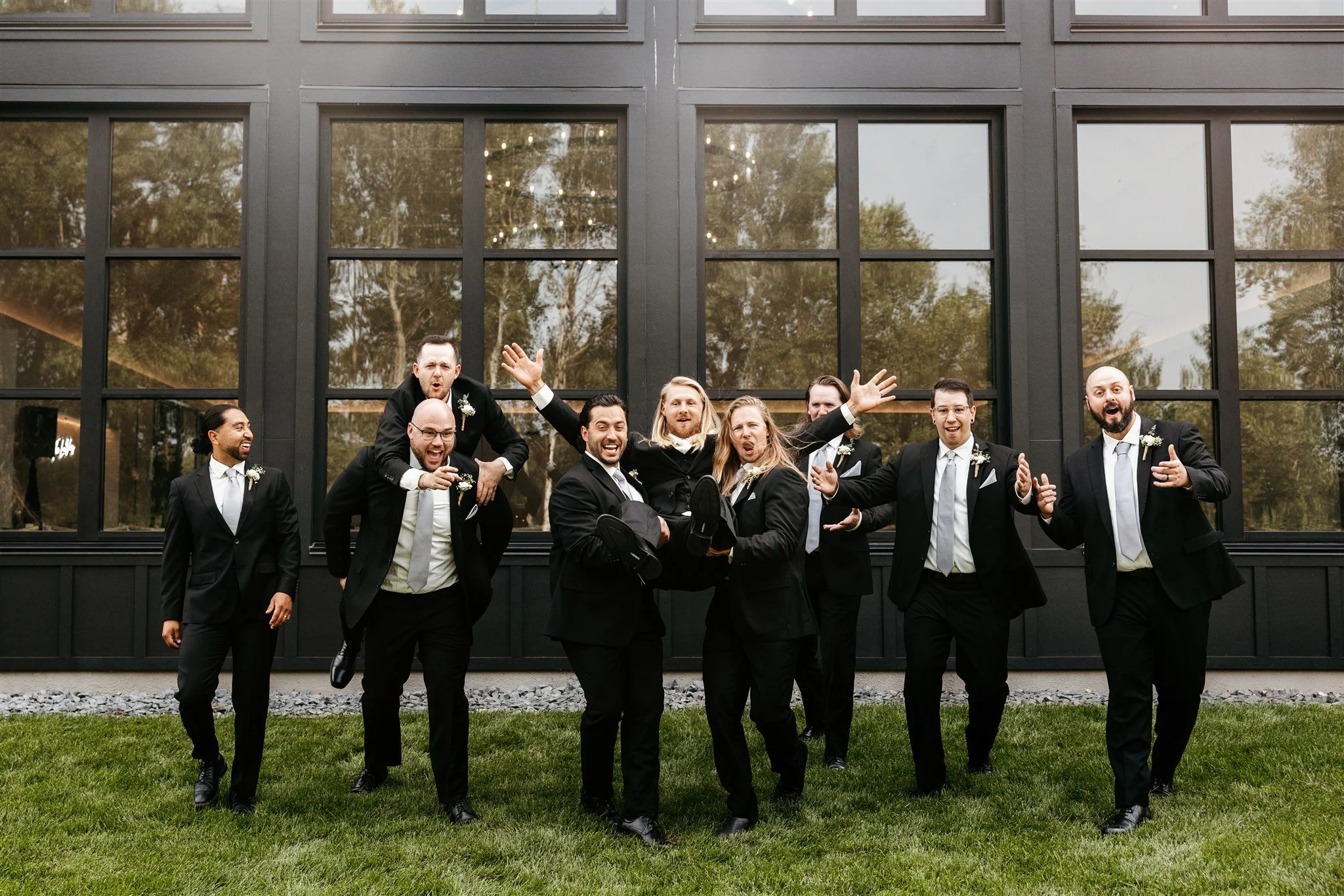 A group of men dressed in tuxedos standing and posing outside in front of a building with large windows, some are smiling and laughing, others making playful gestures.
