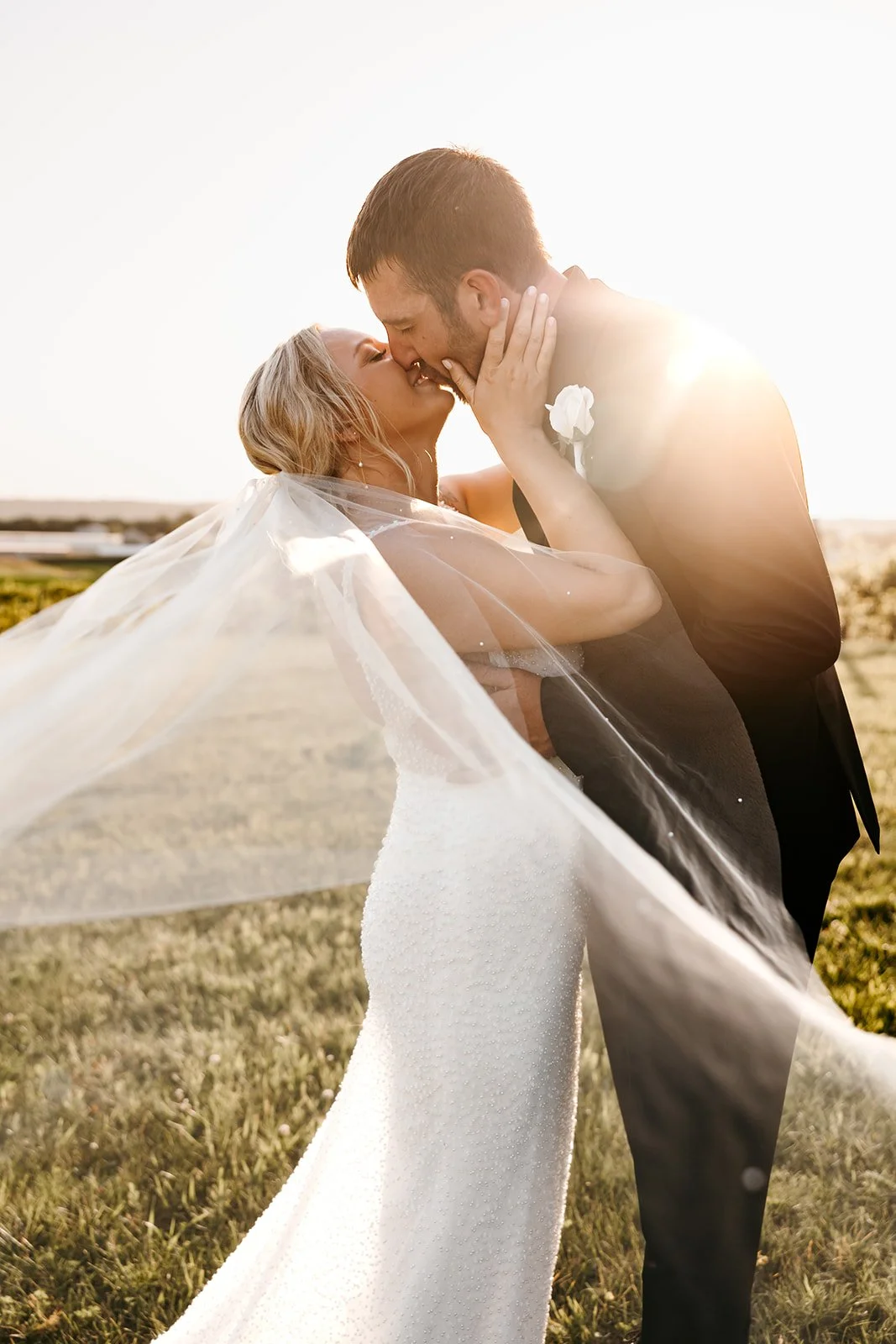 A bride and groom sharing a kiss outdoors on their wedding day at sunset, with the bride holding onto the groom's face, her veil flowing in the wind.