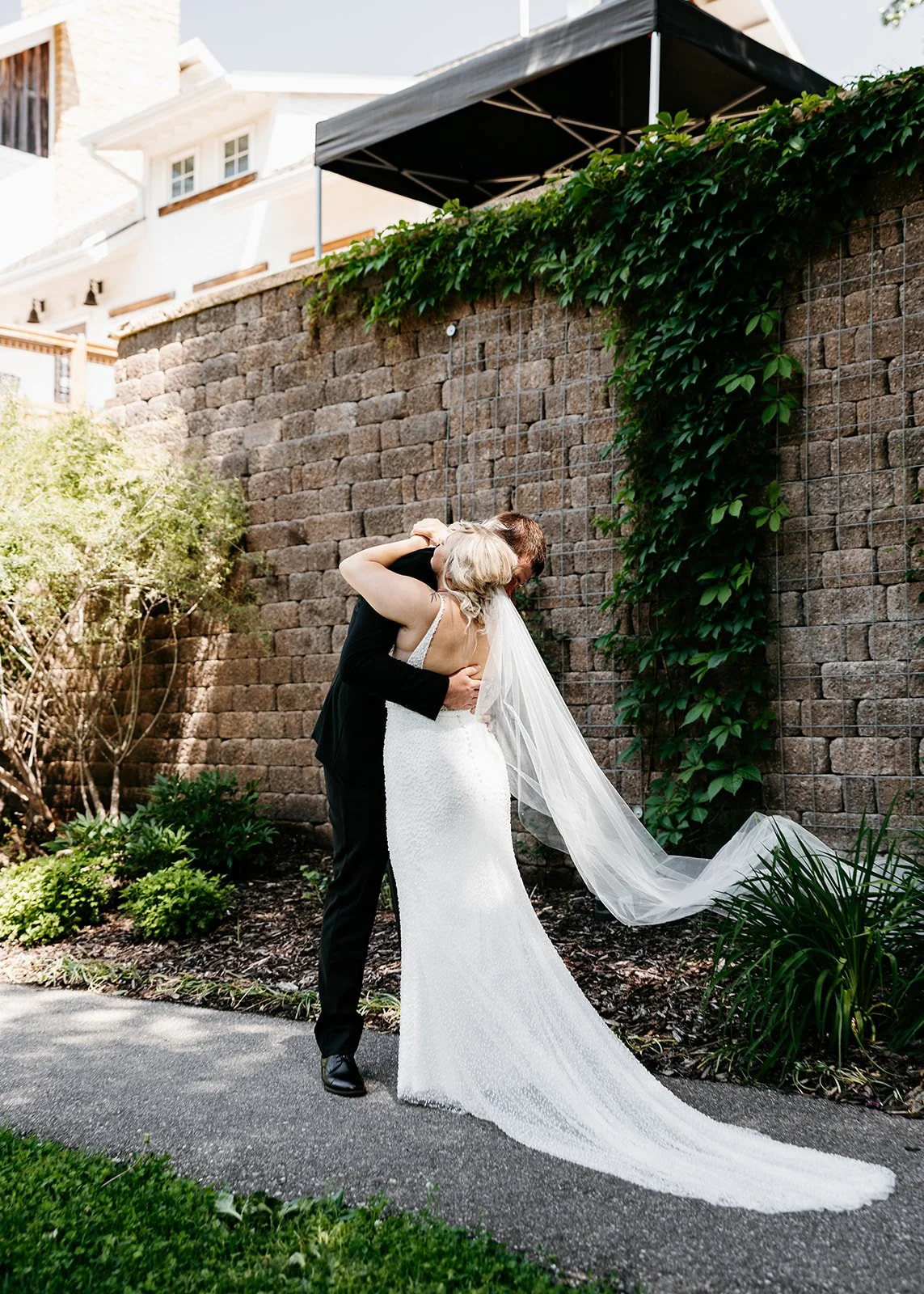 A bride and groom embrace in a romantic outdoor setting with a stone wall and climbing greenery.