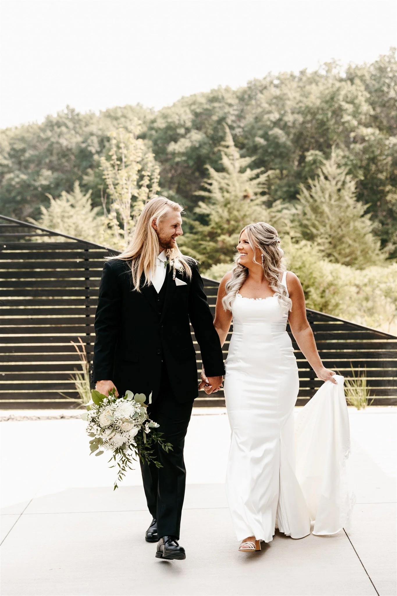 A bride and groom walking hand in hand outdoors, smiling at each other, with the bride holding her wedding dress and the groom holding a bouquet of white flowers, surrounded by greenery.