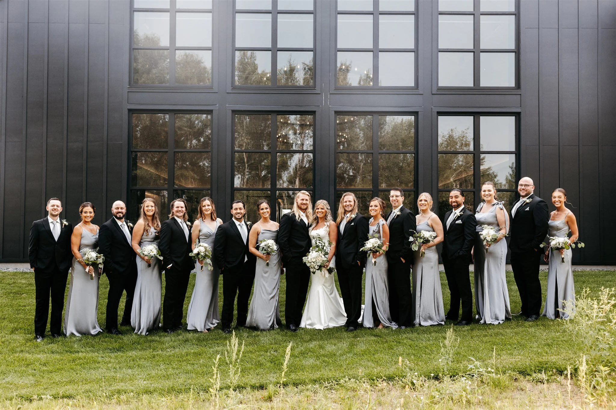 A wedding party of fourteen people, including the bride and groom, posing outside in front of a modern black building with large windows. The bridesmaids are in gray dresses and the groomsmen are in black suits with bow ties. The bride is holding a b