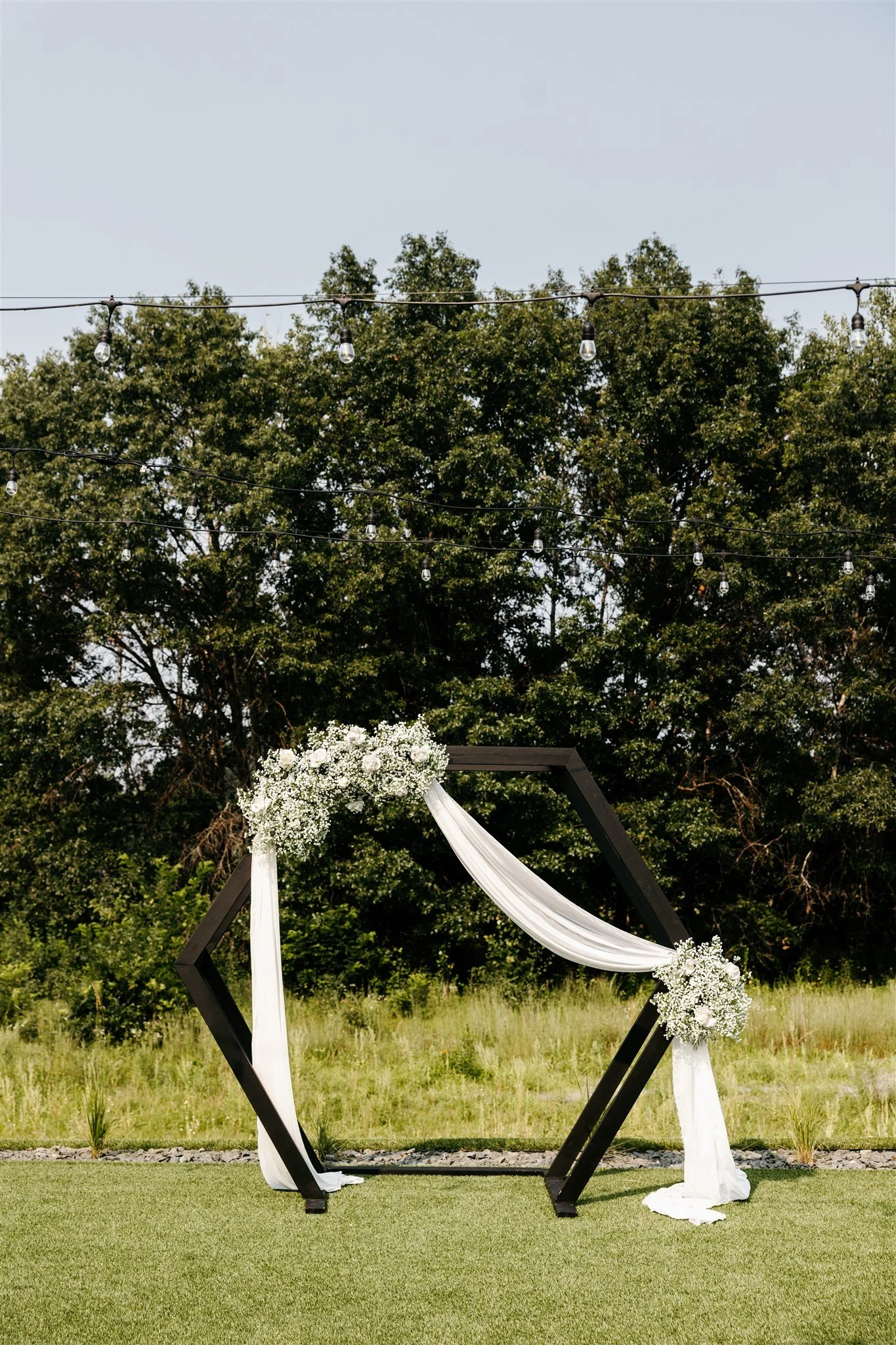 An outdoor wedding arch decorated with white drapery and white flowers, set against a background of green trees, with string lights overhead.