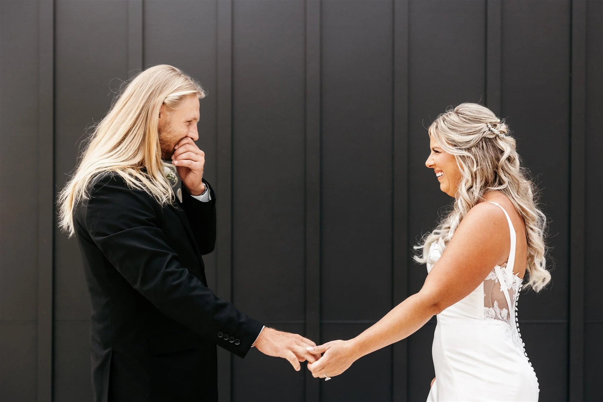 A bride and groom holding hands and smiling at each other during their wedding ceremony, with the groom covering his mouth in joy and the bride showing happiness.