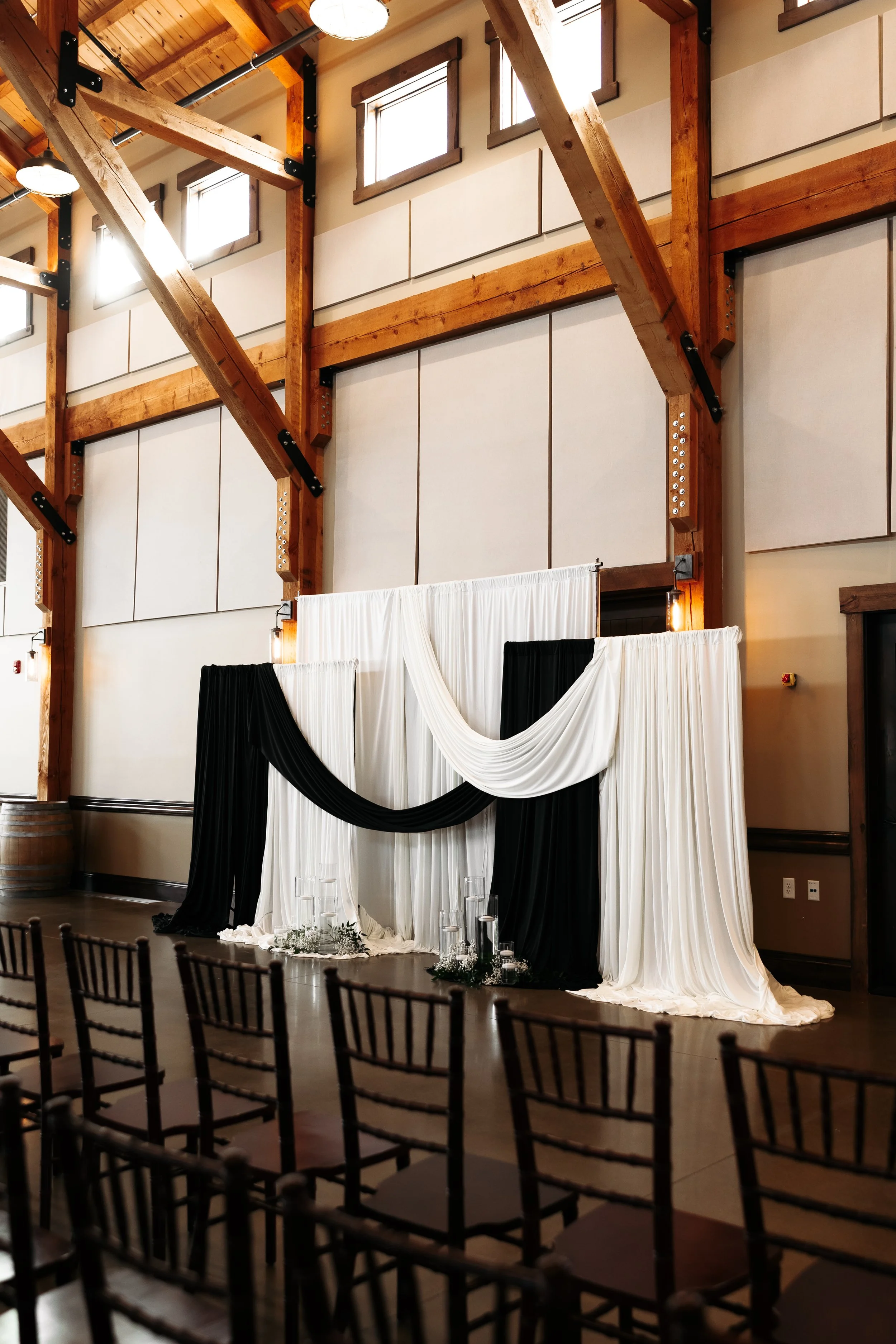 Decorative backdrop with white and black drapery, set up in a rustic event hall with wooden beams and chairs in the foreground for a wedding or special event.