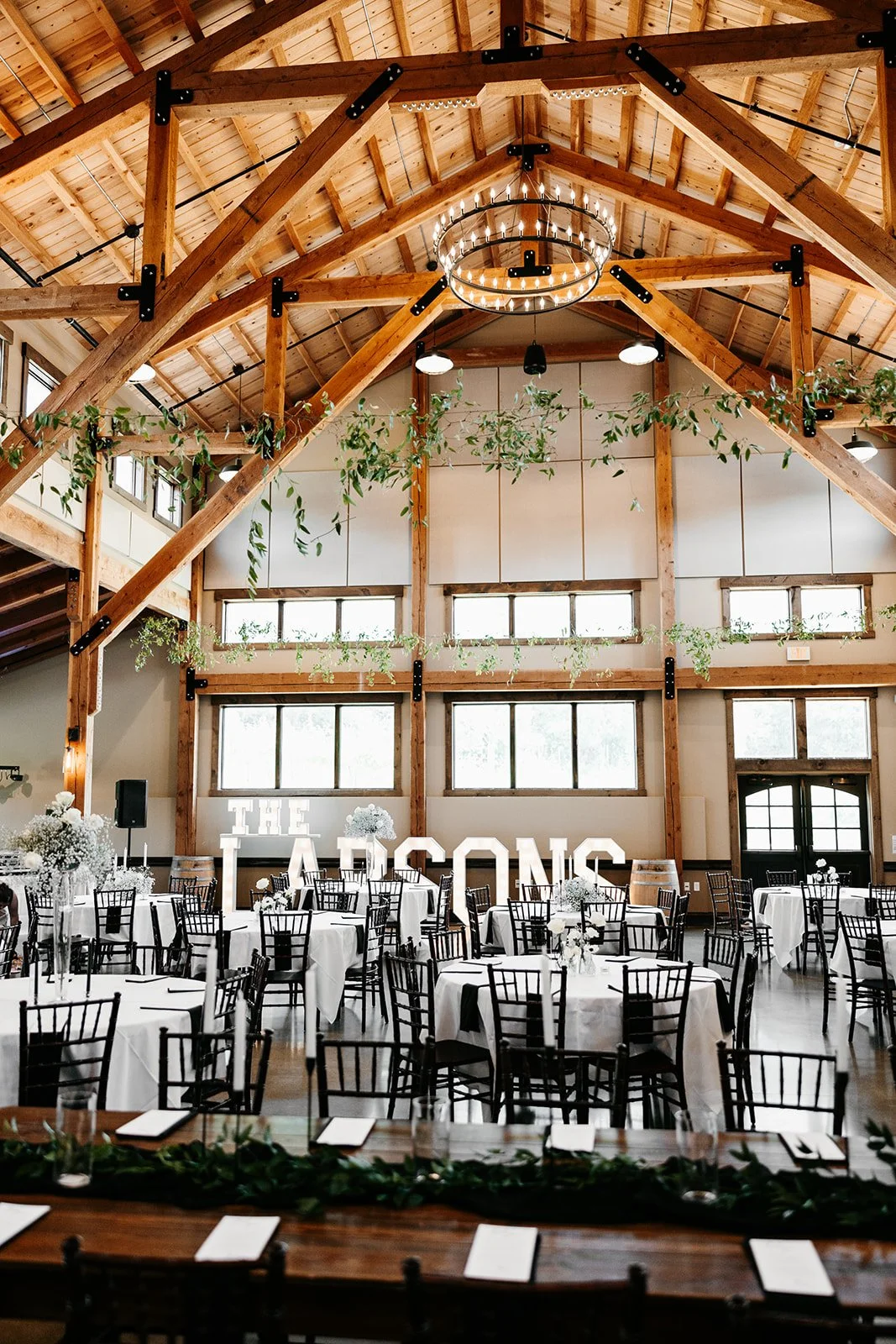 Indoor wedding reception with white tablecloth-covered tables, black chairs, floral centerpieces, and large illuminated letters spelling "THE LADSONS" in the background. The venue has high wooden ceilings, exposed beams, and windows.