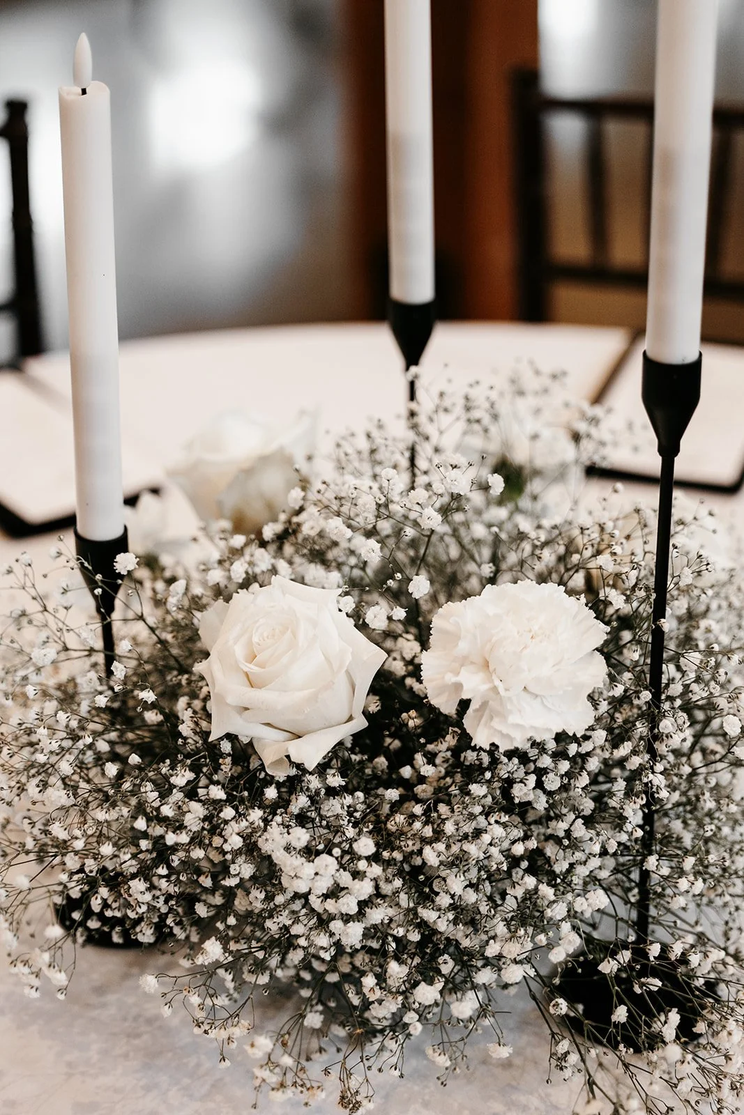 White floral arrangement with roses and baby's breath on a table, surrounded by white taper candles in black candle holders.
