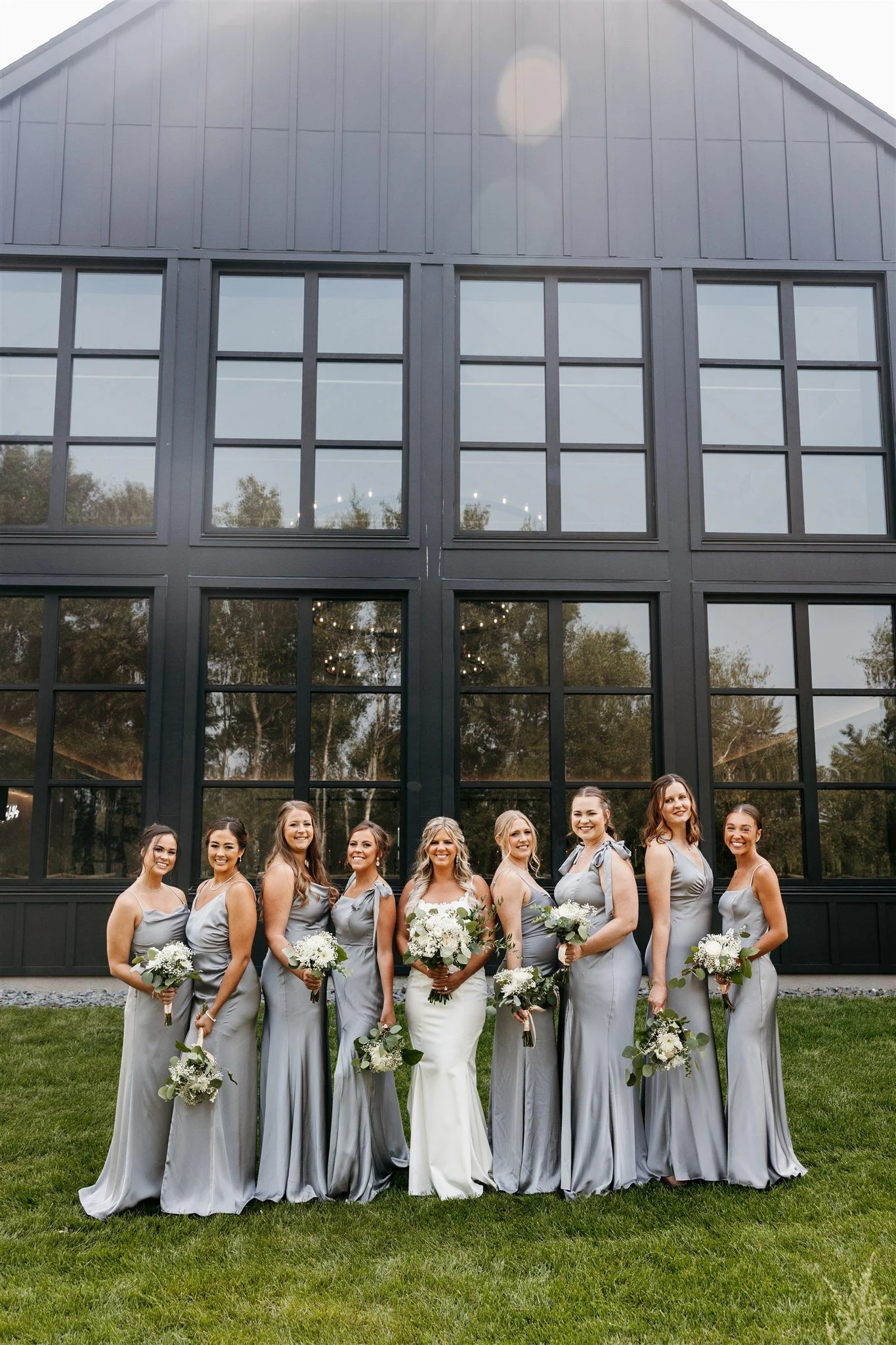 A bride and eight bridesmaids are standing outside in front of a large black building with multiple windows. They are all wearing matching gray dresses and holding bouquets of white flowers.