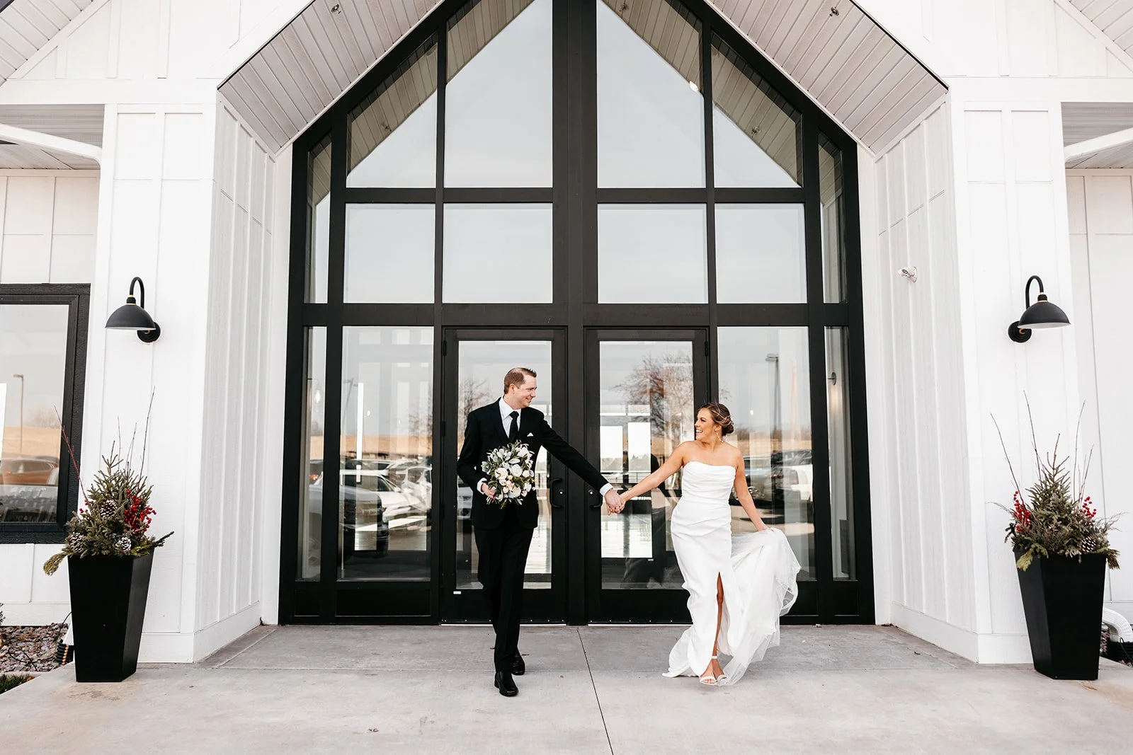 A bride and groom holding hands in front of a modern building with large glass windows, wedding flowers on both sides.