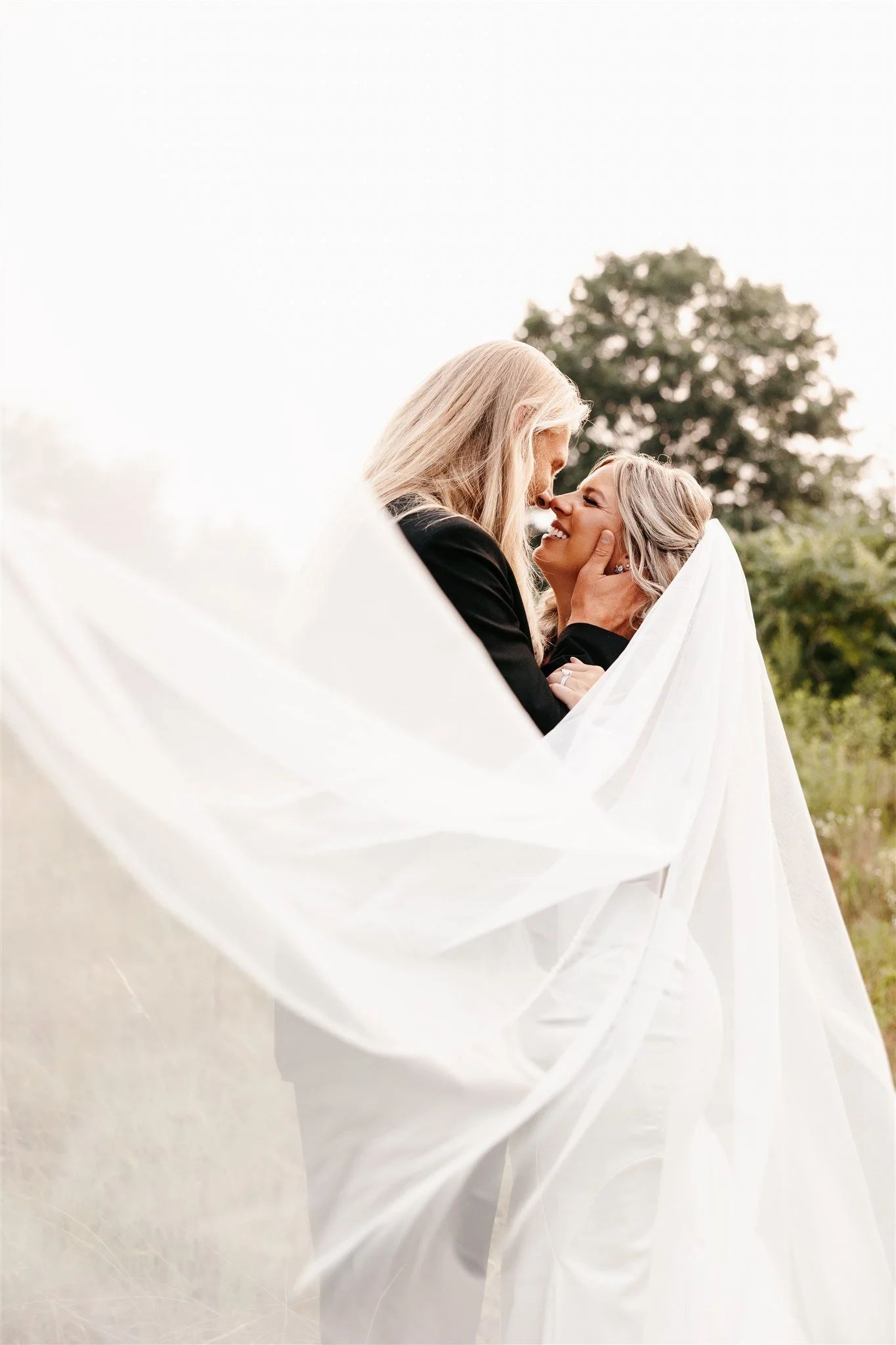 A couple, possibly newlyweds, sharing an intimate moment outdoors. The woman is wrapped in a sheer white fabric, and they are smiling at each other with noses touching, with a tree in the background.