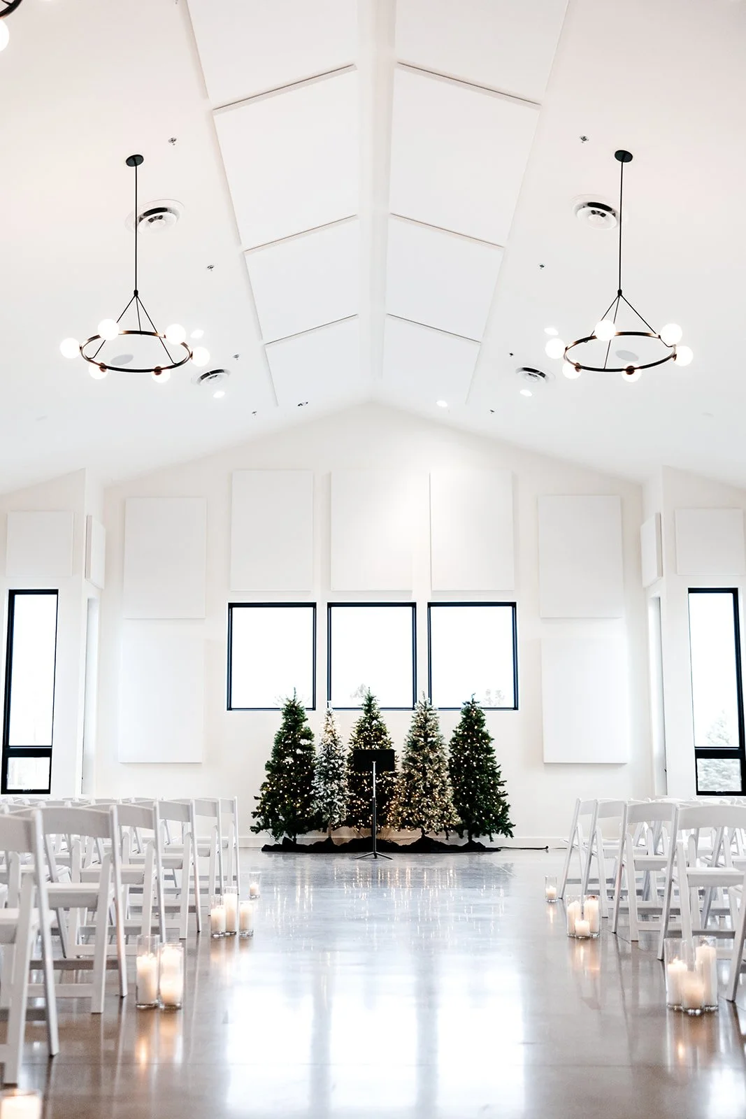 Bright white decorated Christmas trees in front of large windows inside a spacious, minimalistic church or event hall, with white chairs and candles along the aisle for a holiday celebration or wedding.