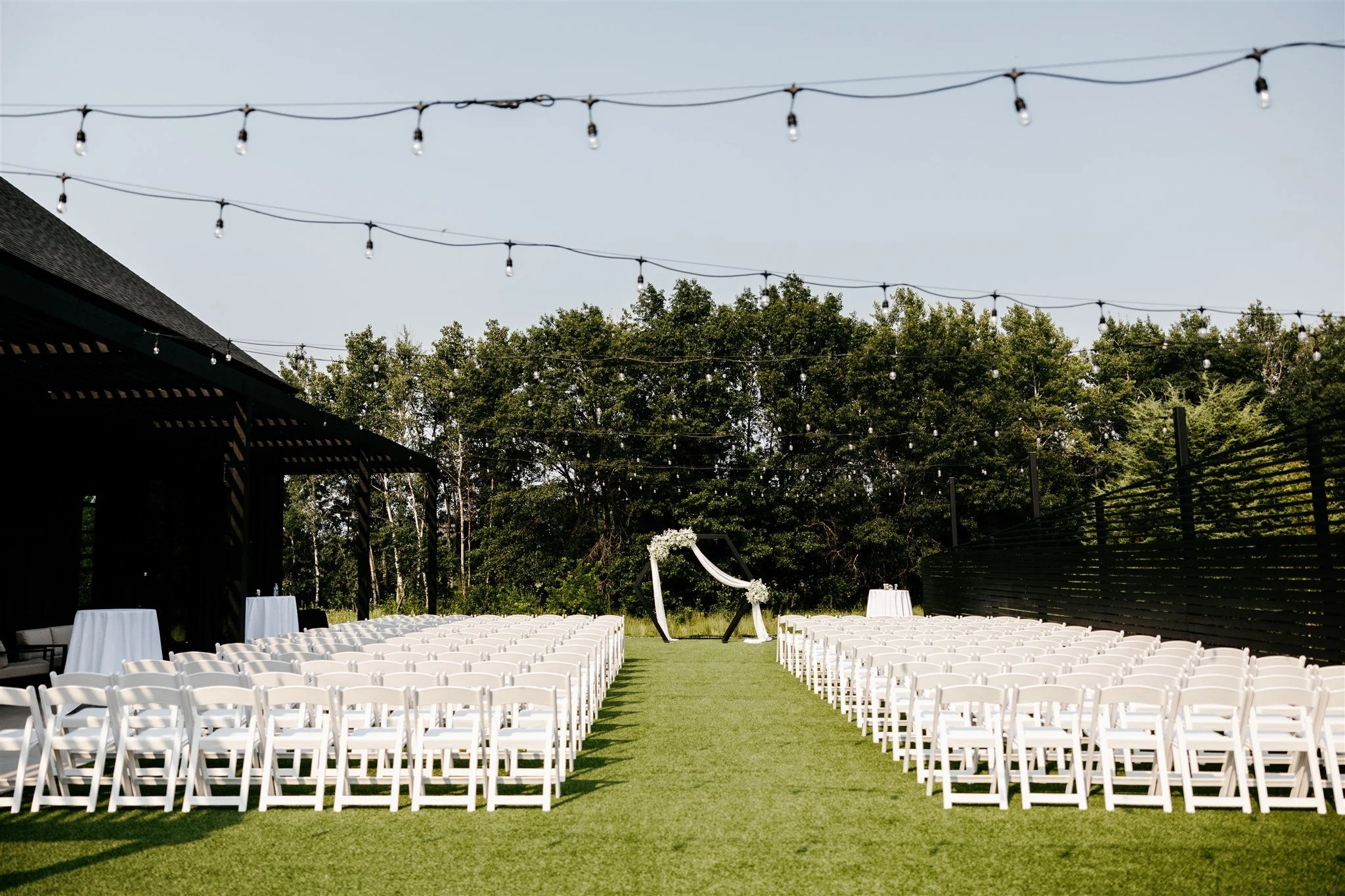 Outdoor wedding setup with white chairs arranged in rows facing a decorated arch on a grassy lawn, string lights overhead, trees in the background.