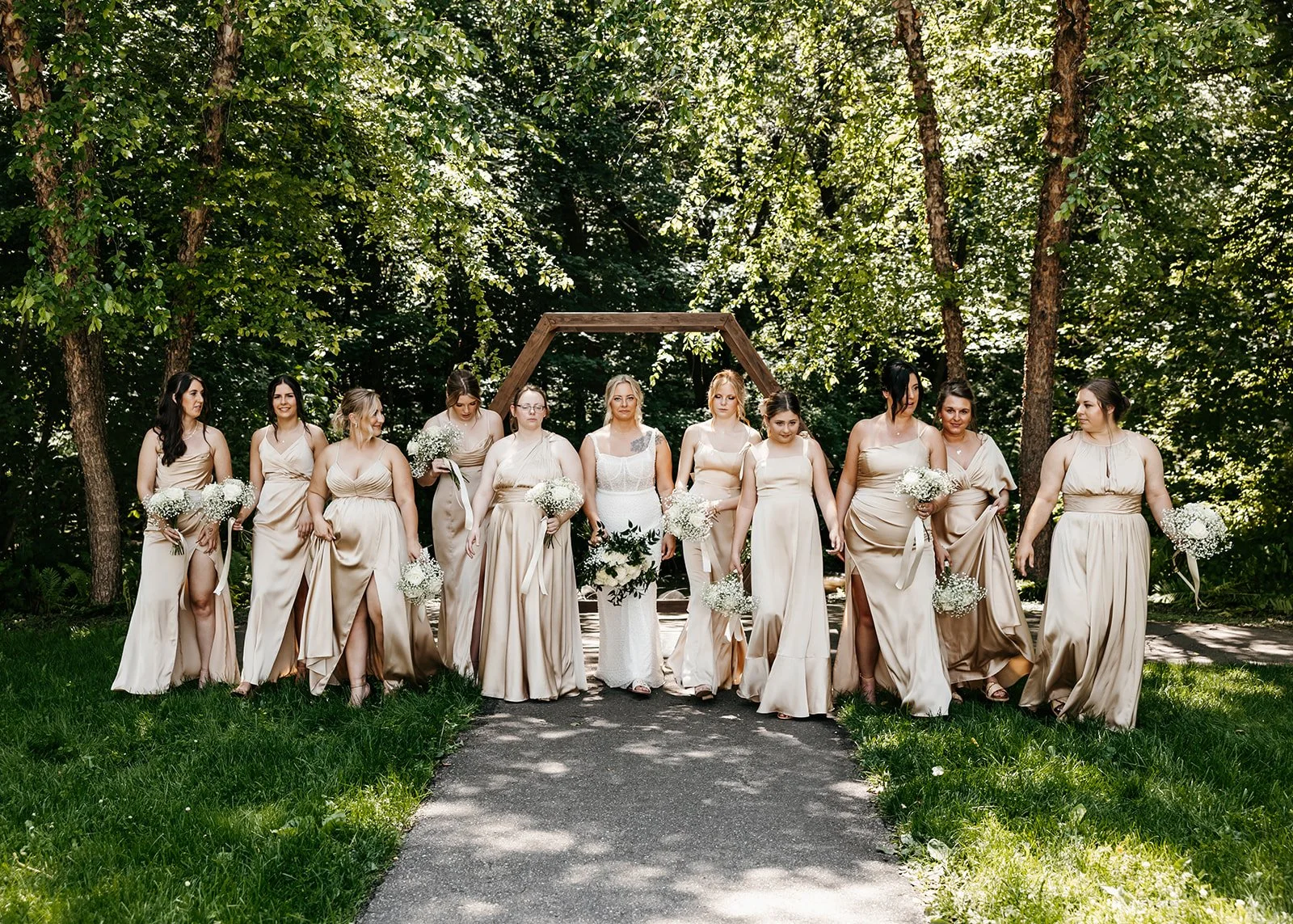 Bride and bridesmaids standing outdoors on a wooded pathway during a wedding photoshoot, holding bouquets of white flowers, under a wooden arch.