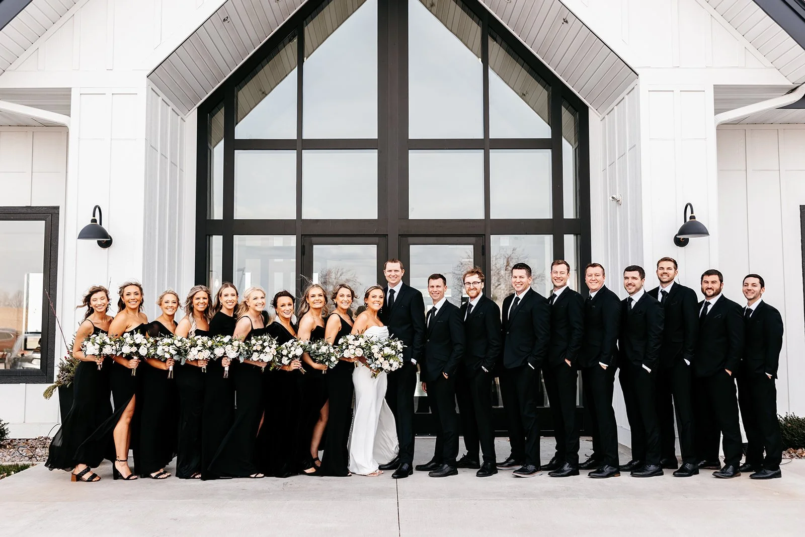 Group of wedding party members dressed in formal attire standing outside in front of a modern building with large glass windows. The women are in black dresses holding bouquets, and the men are in black suits. The bride and groom are center, with the