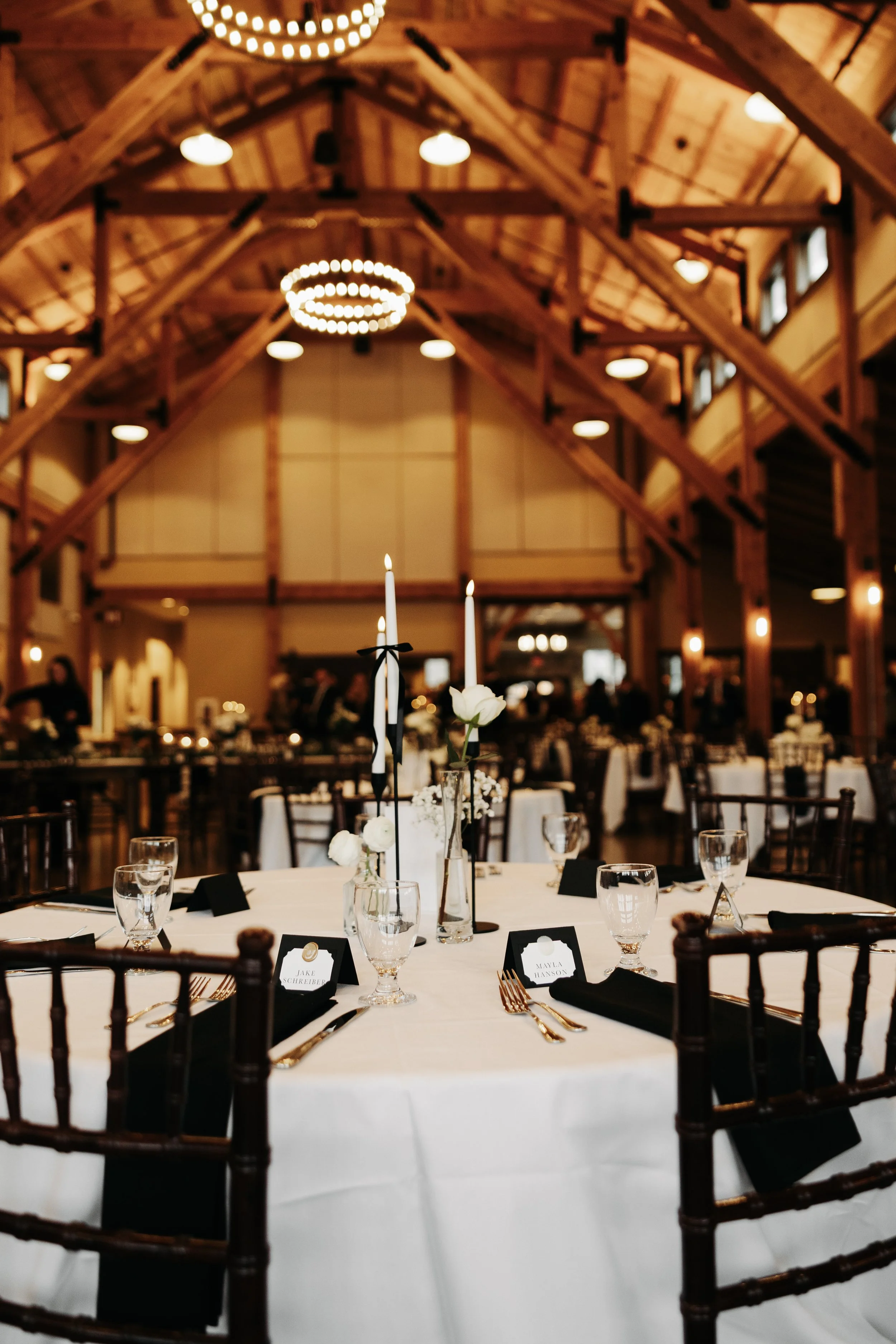 A decorated round dining table set for a formal event with black napkins, gold utensils, water glasses, and small black name cards, featuring a centerpiece with white flowers and tall white candles, inside a rustic wooden-structured banquet hall with