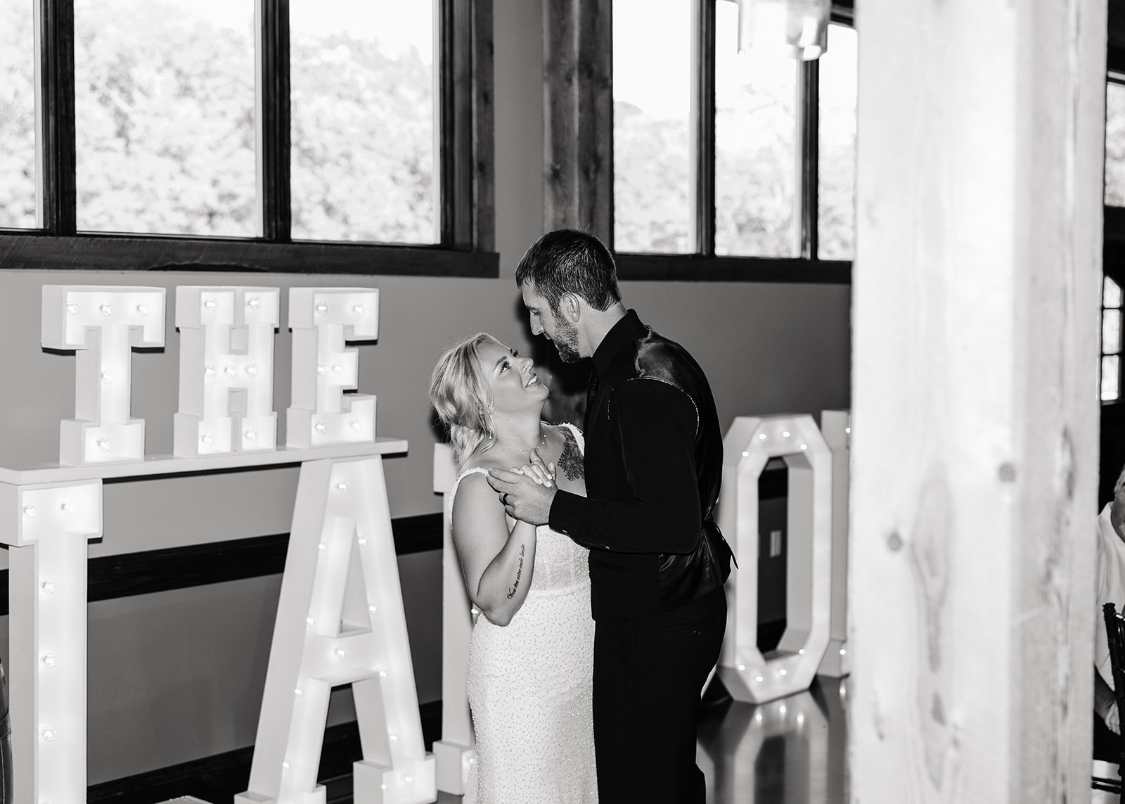 A couple dancing at a wedding reception, with large illuminated letters spelling "THE A" in the background, inside a venue with large windows.