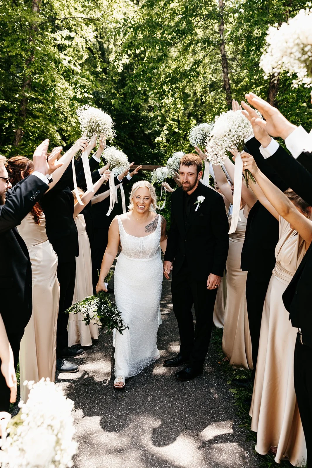 A newlywed couple walking through a floral arch at an outdoor wedding ceremony, with bridesmaids and groomsmen on either side holding white flower bouquets, surrounded by lush green trees.