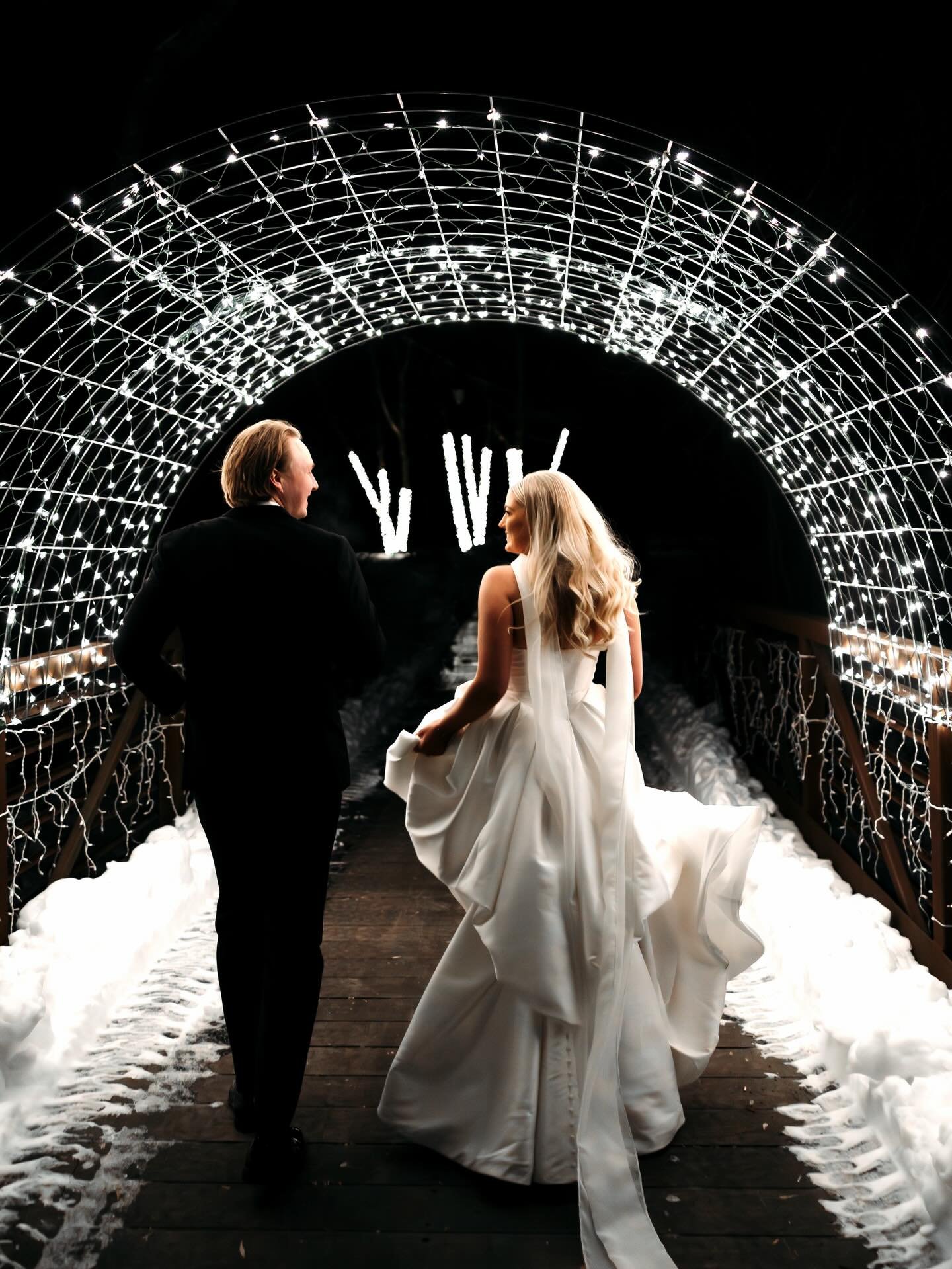 A bride and groom walking hand in hand on a snowy wooden bridge decorated with bright white holiday lights in a tunnel, at night.