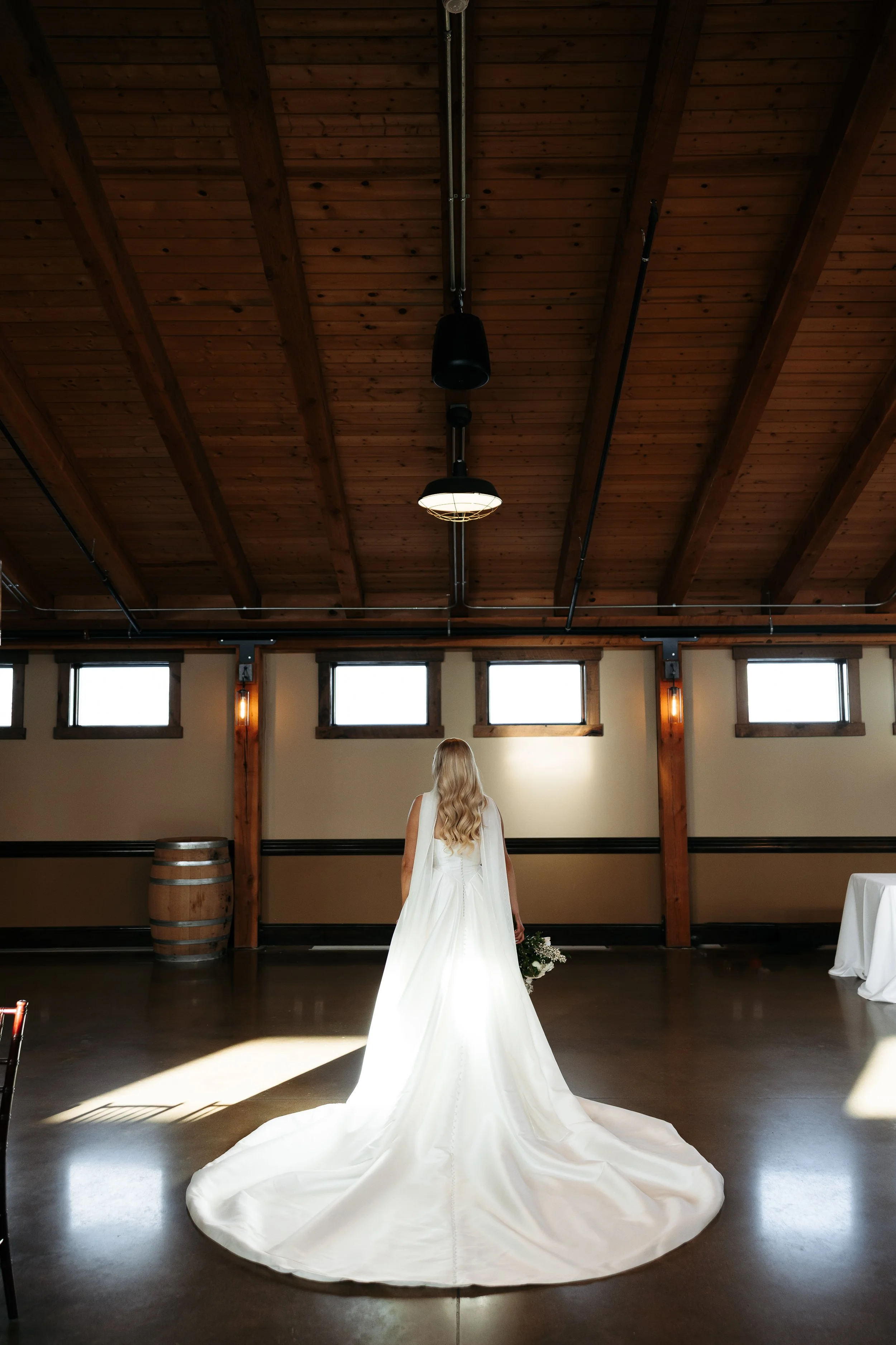 Bride in a white wedding dress holding a bouquet, standing alone in an indoor venue with wooden ceiling and beams, illuminated by natural light through windows.