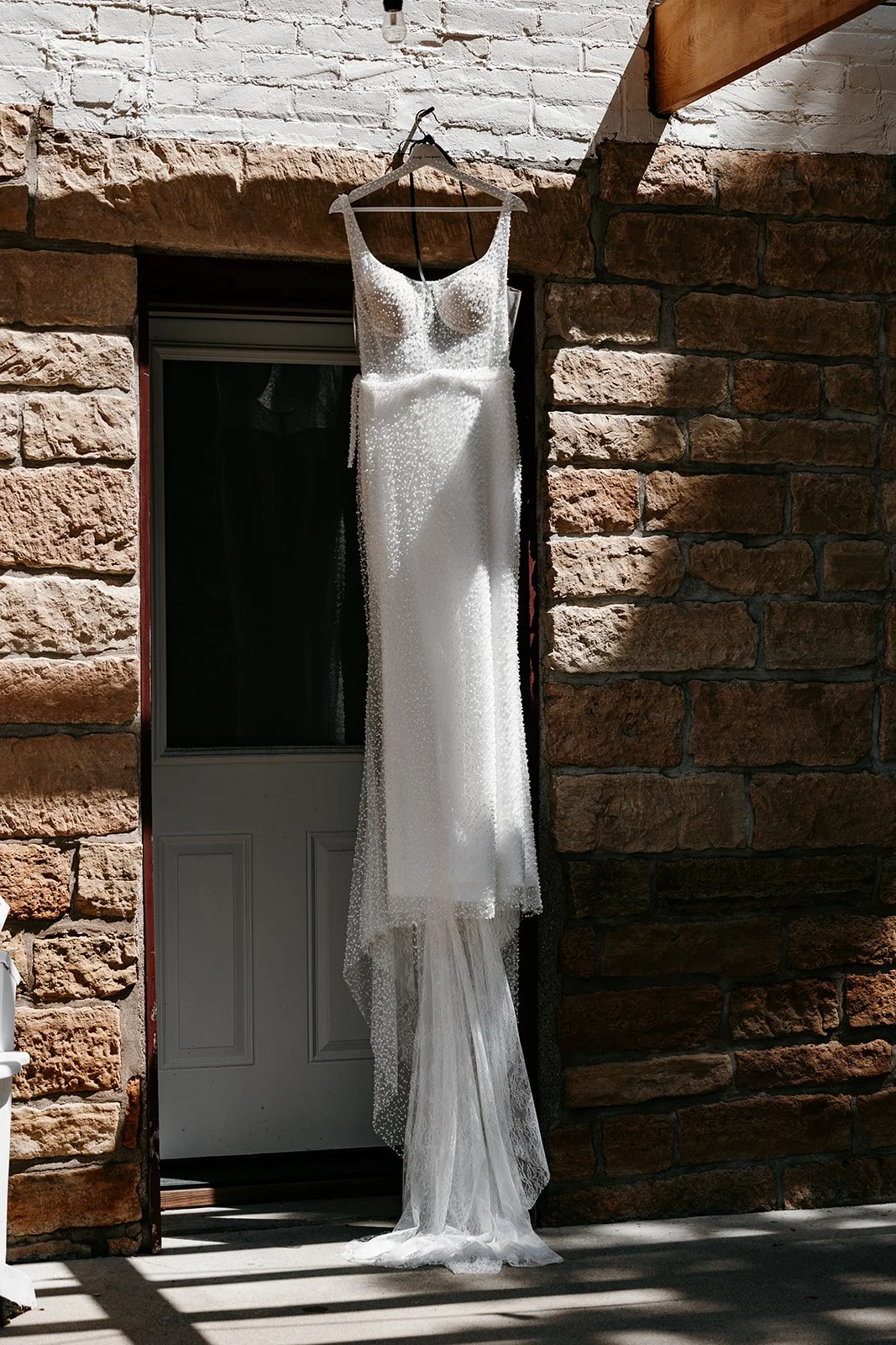 A wedding dress with sparkly embellishments hanging on a hanger outside a brick building, with sunlight and shadows on the ground.