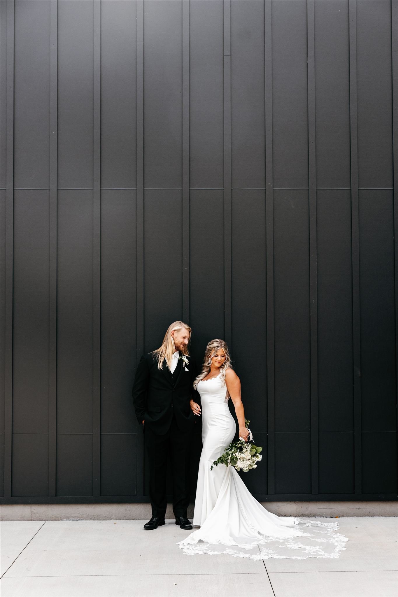 Bride and groom standing against a black wall, with the bride holding a bouquet of white flowers and wearing a white gown, and the groom dressed in a black suit.