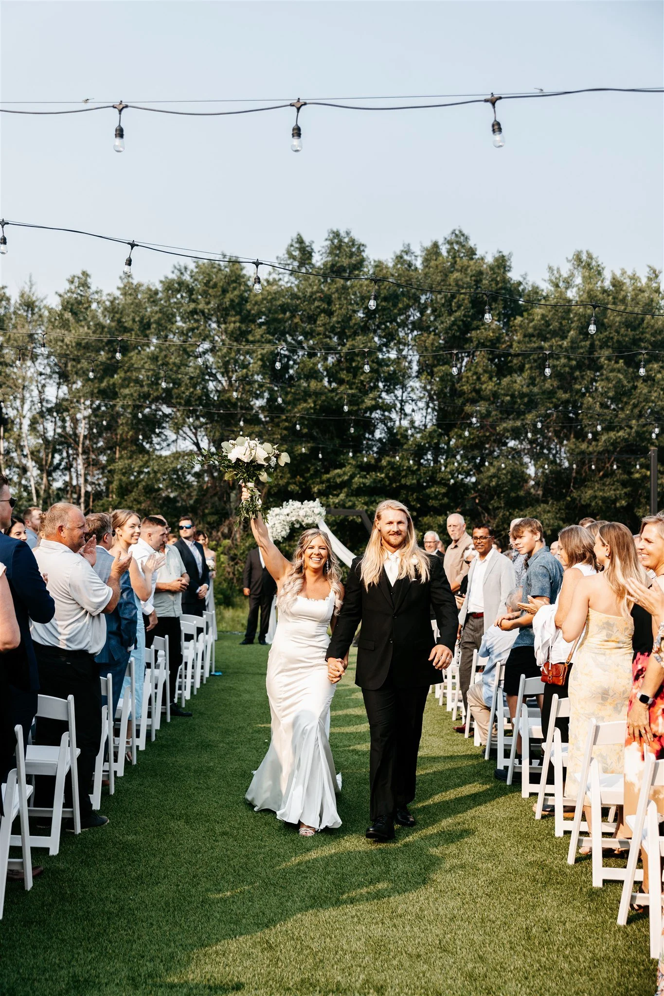 A newlywed couple walking down the aisle at an outdoor wedding ceremony, with guests standing and clapping on either side, under string lights on a sunny day.