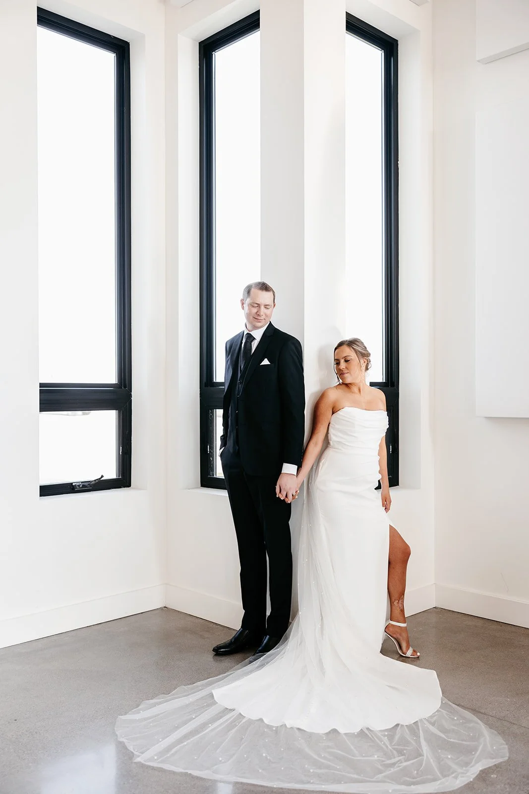 A bride and groom hold hands in front of large windows, with the bride wearing a strapless white wedding gown and the groom in a black suit, in a bright, modern room.