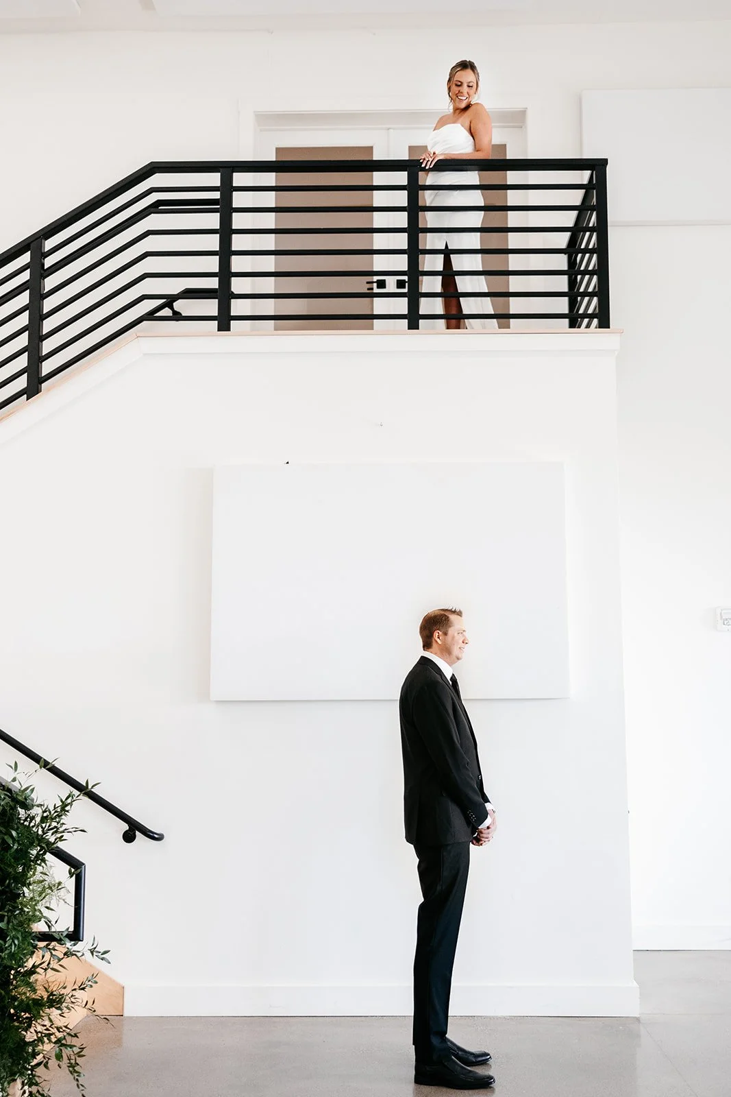A woman in a white dress standing on an upper balcony and smiling, facing a man in a black suit standing on the ground floor in a bright, minimalist interior.