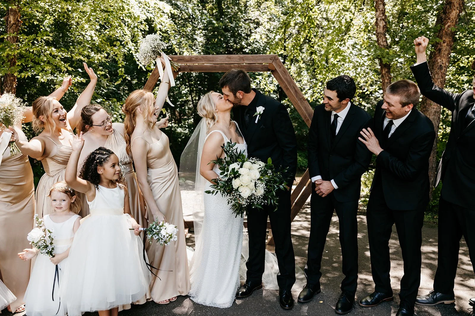 A wedding ceremony outdoors with a bride and groom kissing under a wooden arch, surrounded by bridesmaids and groomsmen, with children holding flowers, all celebrating.