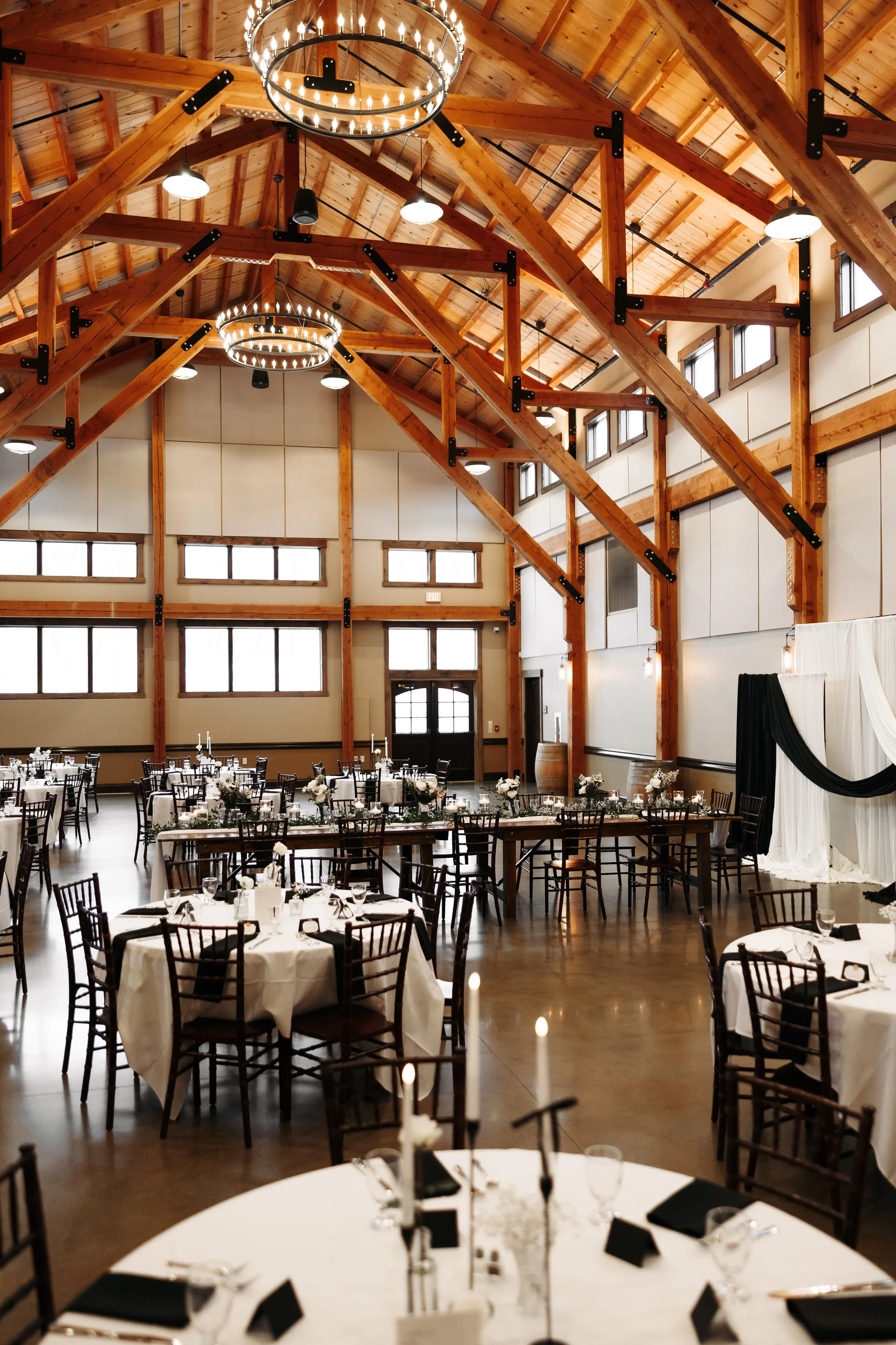 Elegant wedding reception hall with round tables covered in white tablecloths and black napkins, decorated with floral centerpieces and candles, in a rustic barn-style venue with wooden beams and large windows.