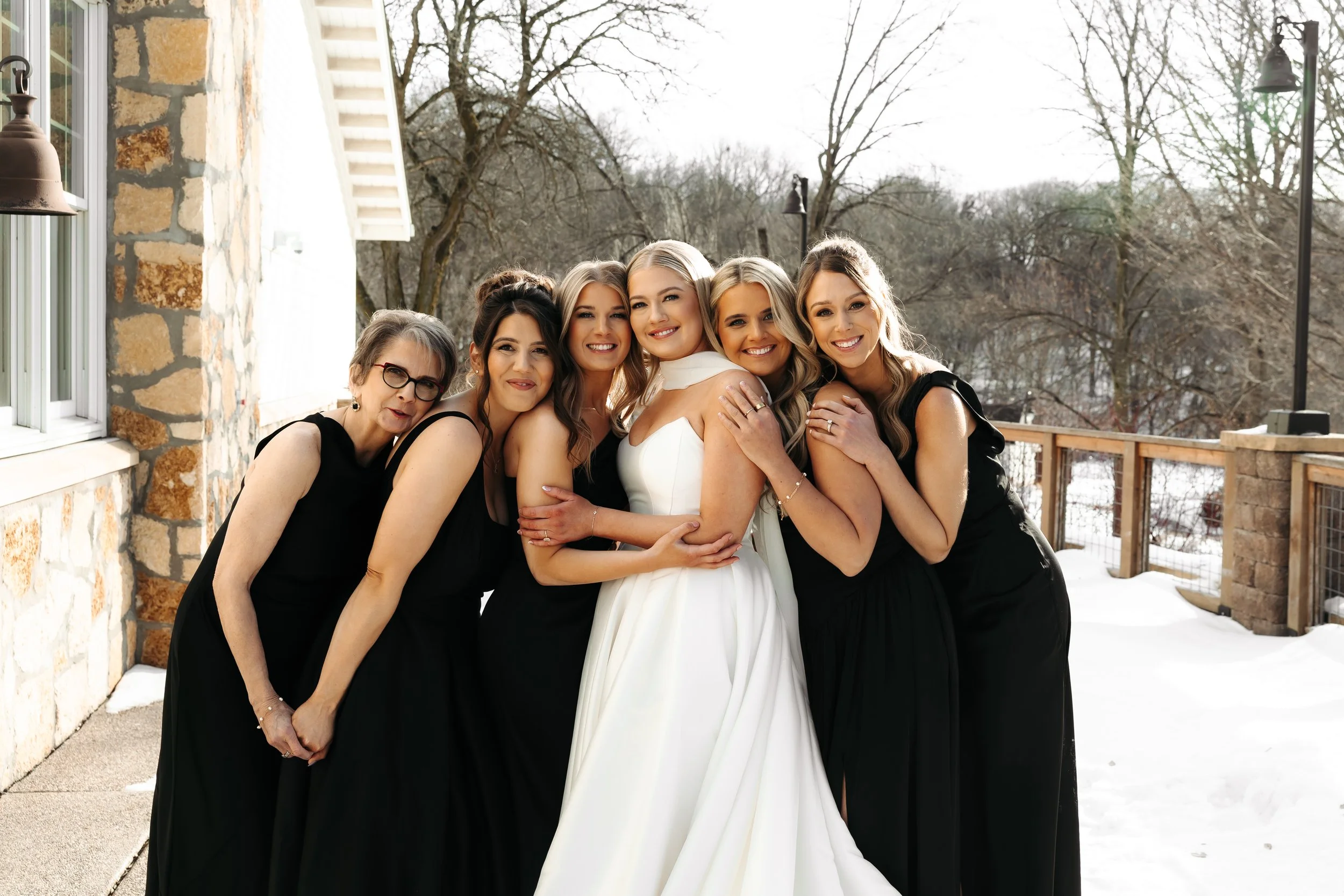 A bride in a white wedding dress surrounded by five women in black dresses, standing on a snowy outdoor porch with trees in the background.
