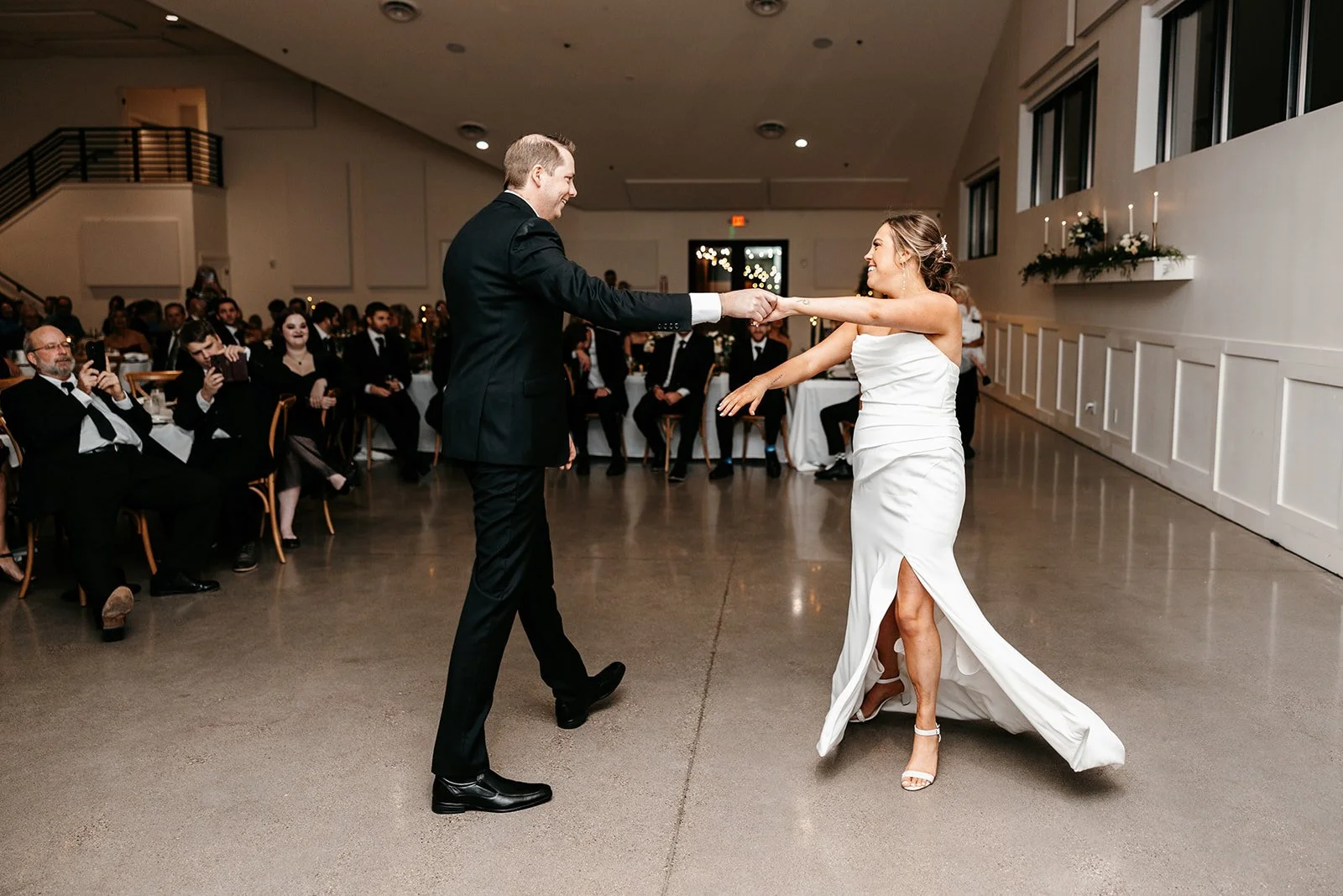A bride and groom dancing together at their wedding reception, holding hands and smiling, with guests watching in the background.