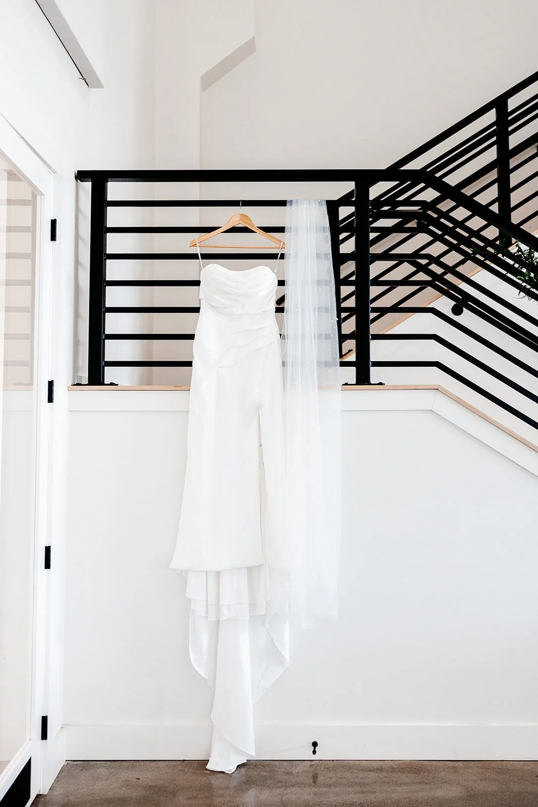 White wedding dress hanging on a wooden hanger on a black railing in a modern, minimal interior.