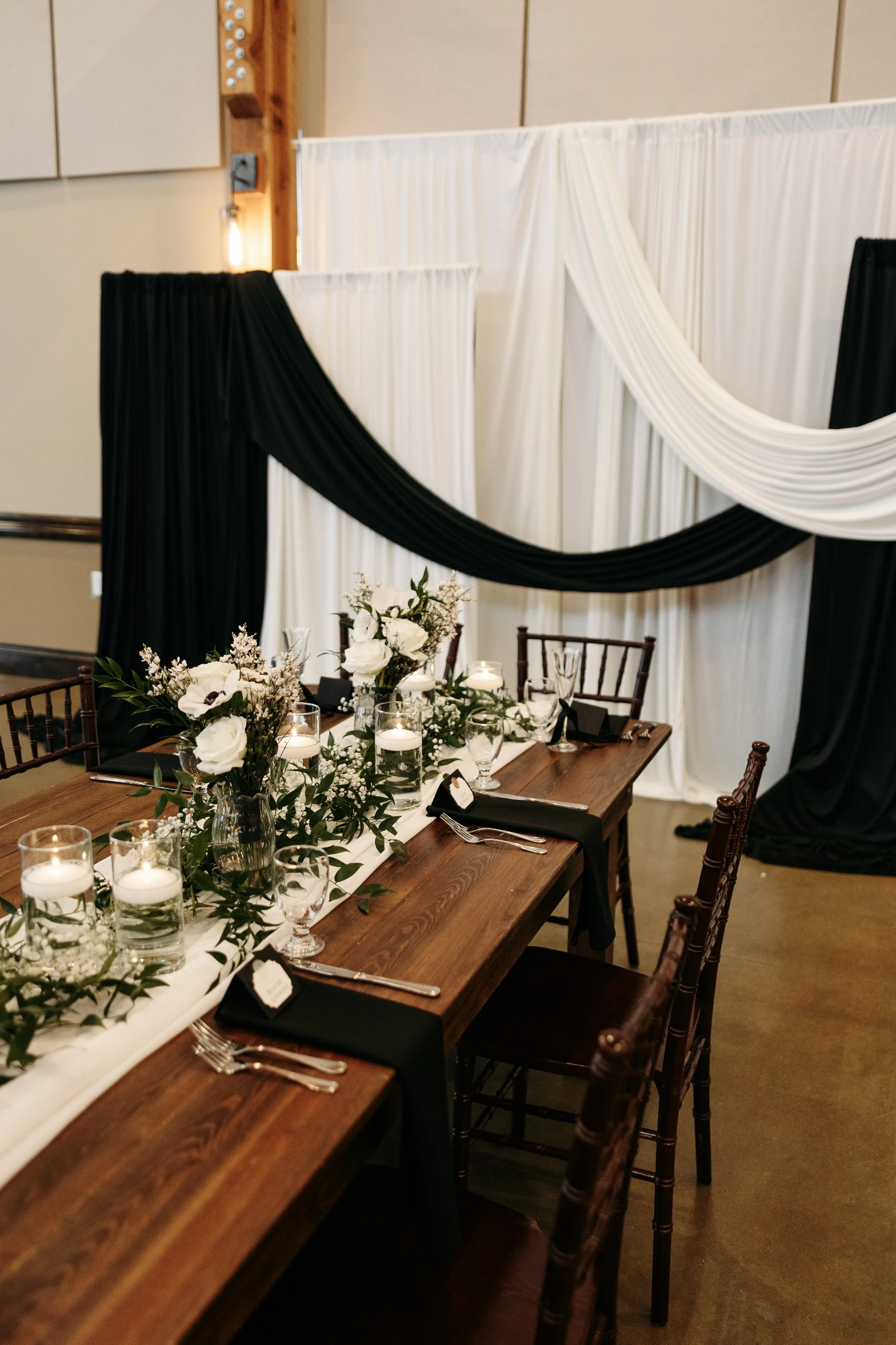 A decorated wedding or event reception table with white and black drapery backdrop.