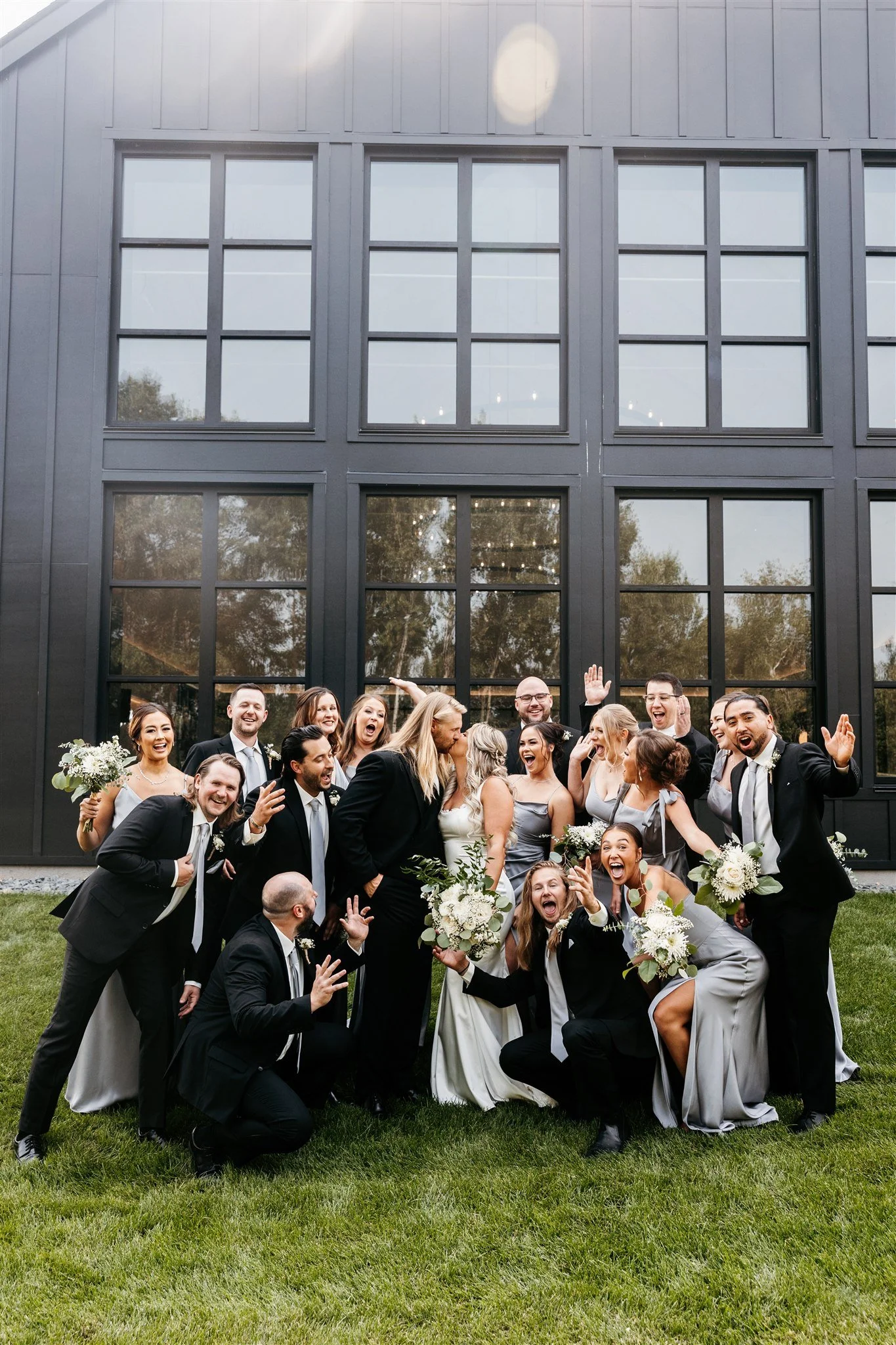 A large group of wedding guests, dressed in formal attire, celebrating outdoors in front of a modern black building with large windows. They are smiling, laughing, and posing for the photo, with some holding bouquets of flowers.