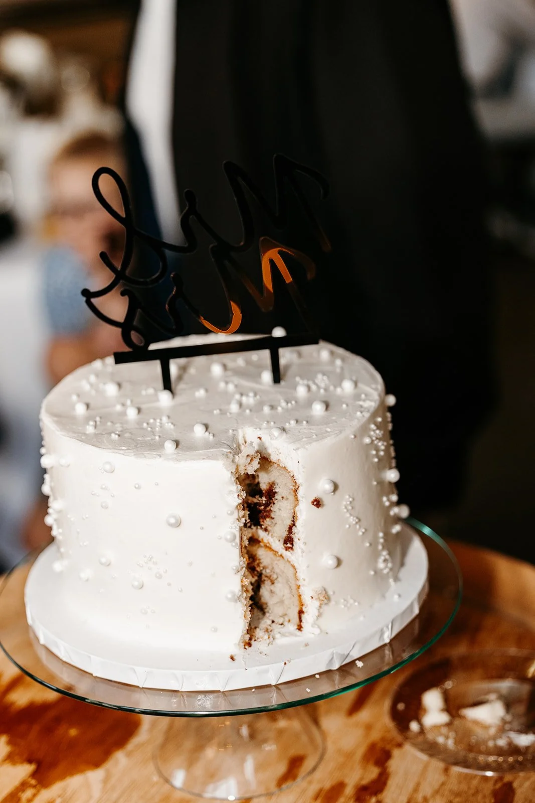 A white cake with pearl decorations and a baked inside, topped with a black and orange 'Happy New Year' cake topper, on a glass cake stand. Part of the cake has been cut, revealing the inside layers.