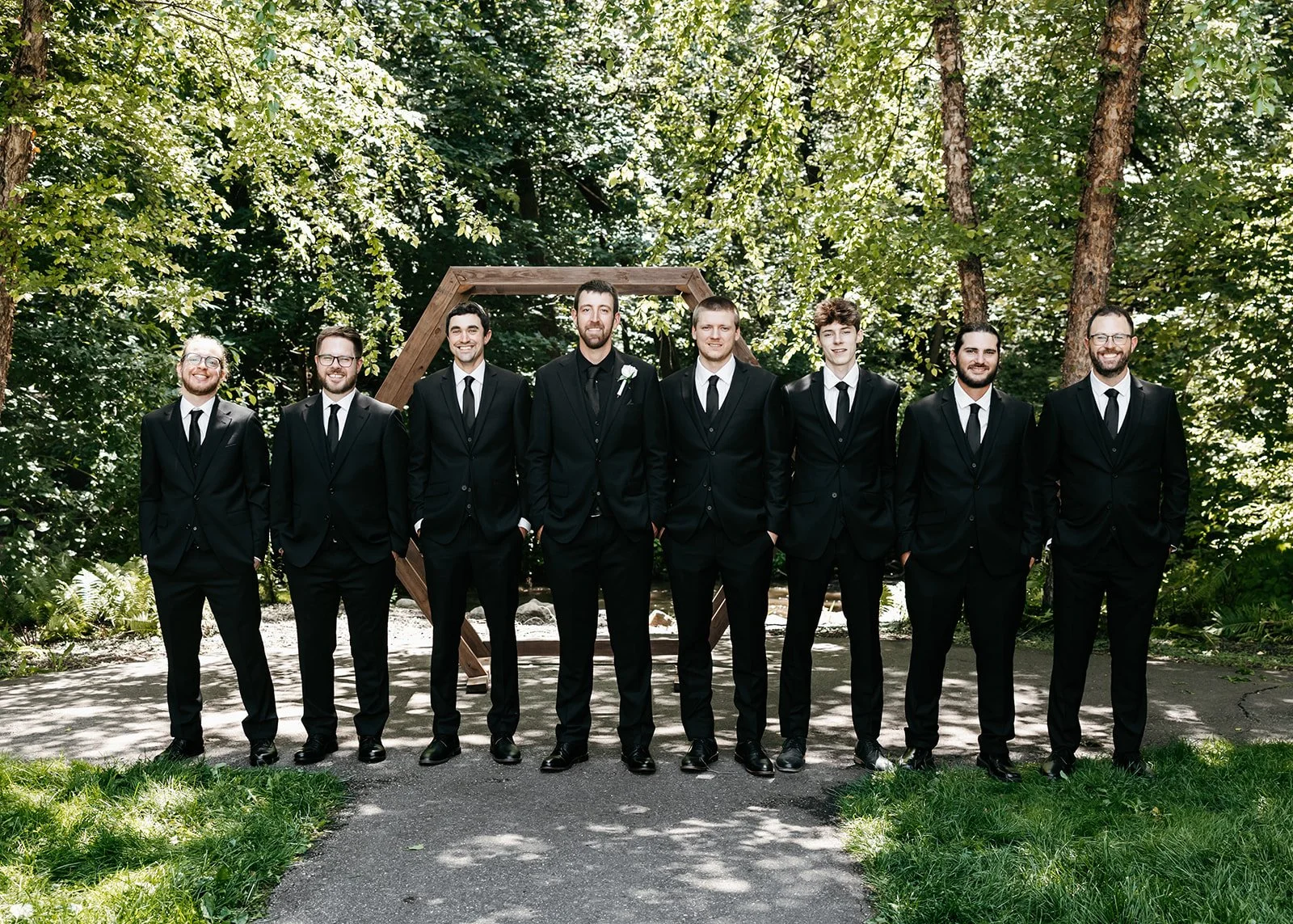 A group of eight men in black suits and ties standing outdoors on a paved pathway in front of a wooden arch, surrounded by green trees and foliage.