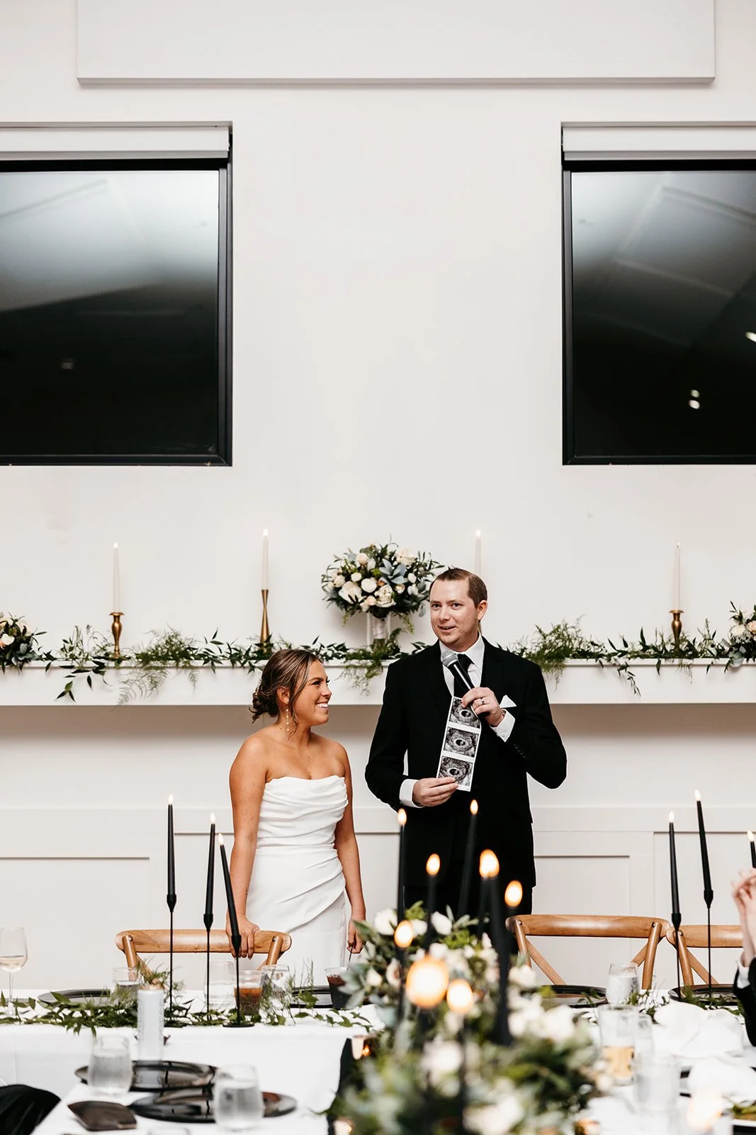Bride and groom standing at a wedding reception, the bride smiling at the groom, who is holding a microphone and a sheet of photos, with candles and floral decorations on the table.